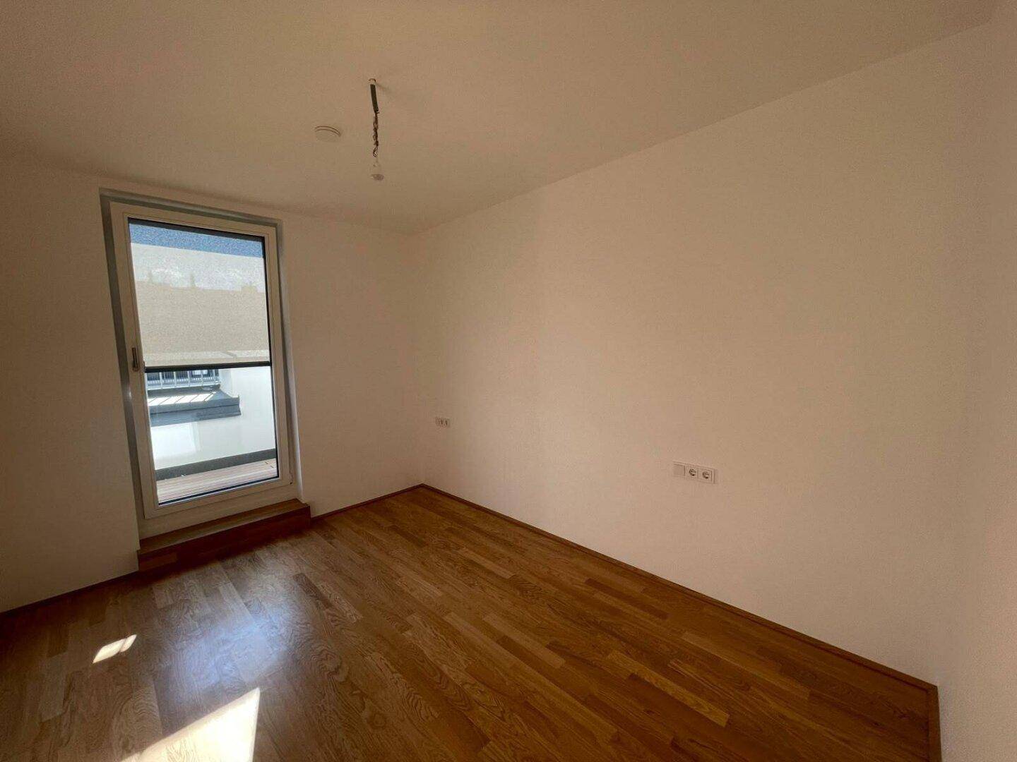 Ruhiges Zimmer mit hellem Parkettboden und einem Fenster, das viel Tageslicht in den Raum bringt.