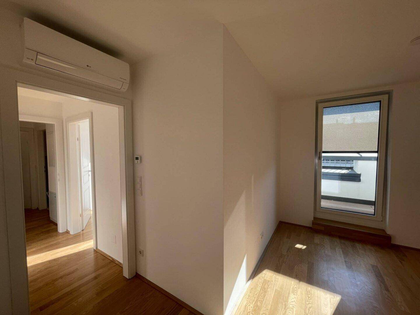 Zimmer mit Parkettboden und Klimaanlage über dem Türrahmen, das zu einem hellen Flur führt.