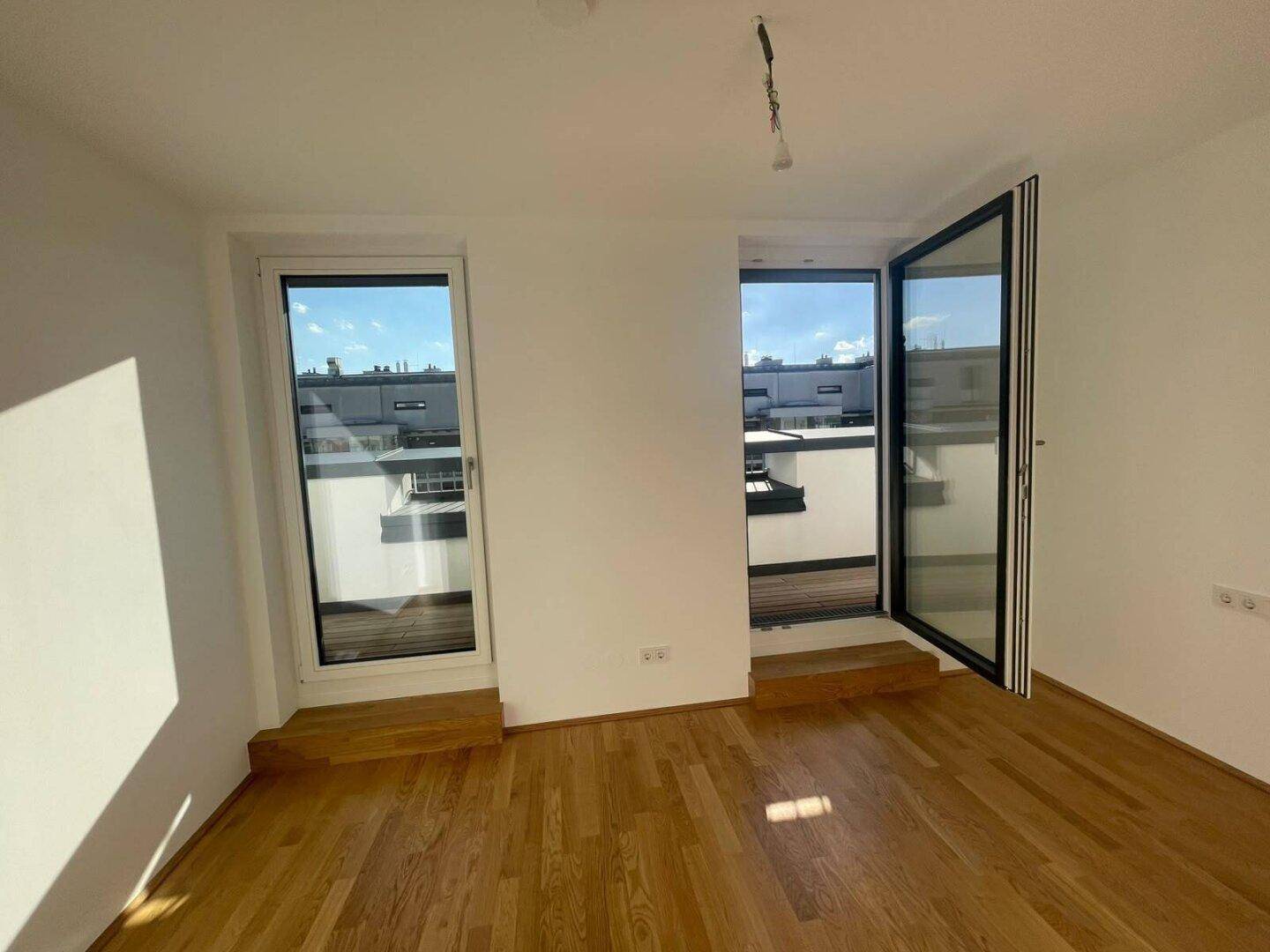 Helles Zimmer mit Parkettboden und zwei großen Fenstern, die Zugang zu einem Außenbereich bieten.