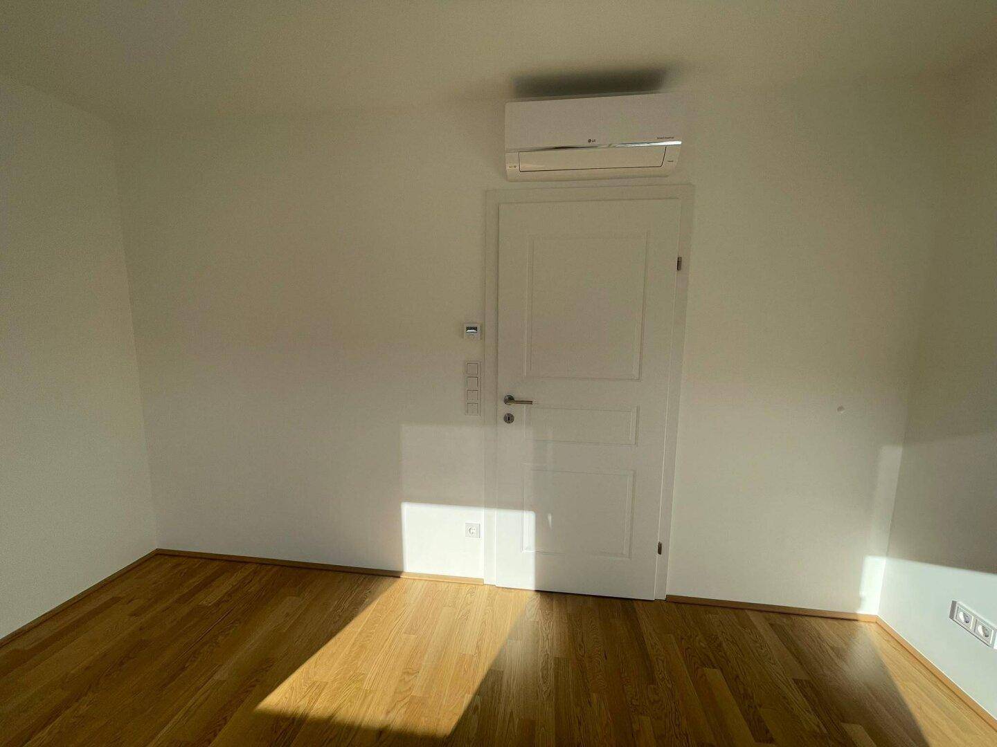 Zimmer mit Parkettboden und Klimaanlage über der Tür, das viel Sonnenlicht durch die Fenster erhält.