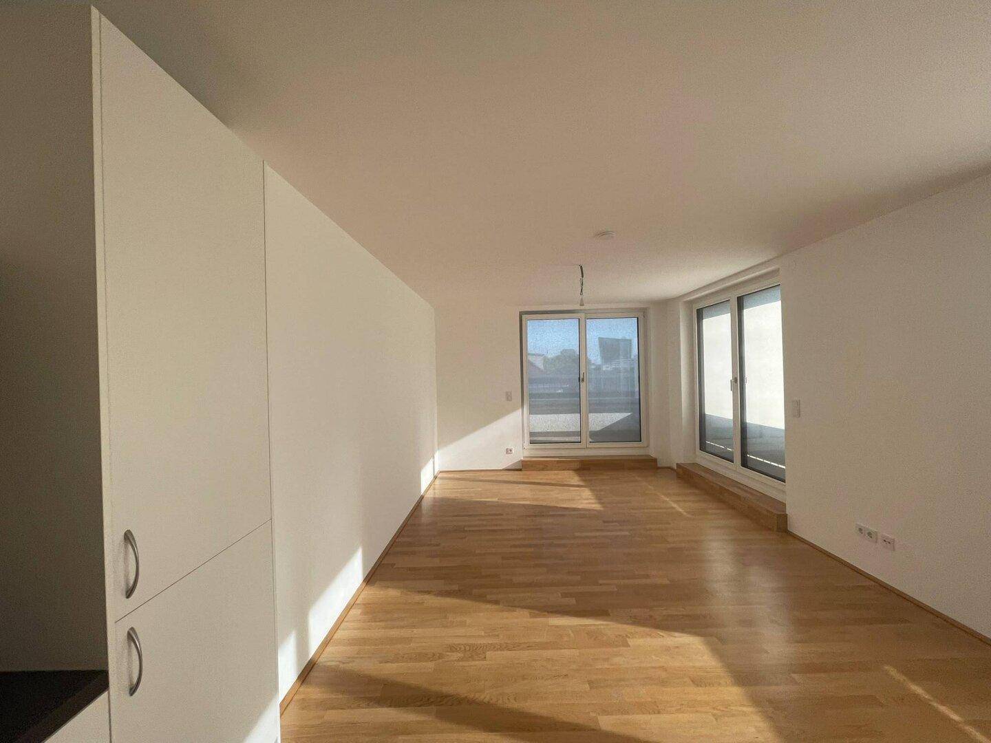 Heller, leerer Wohnbereich mit großen Fenstern, die viel Sonnenlicht hereinlassen, und schönem Parkettboden.