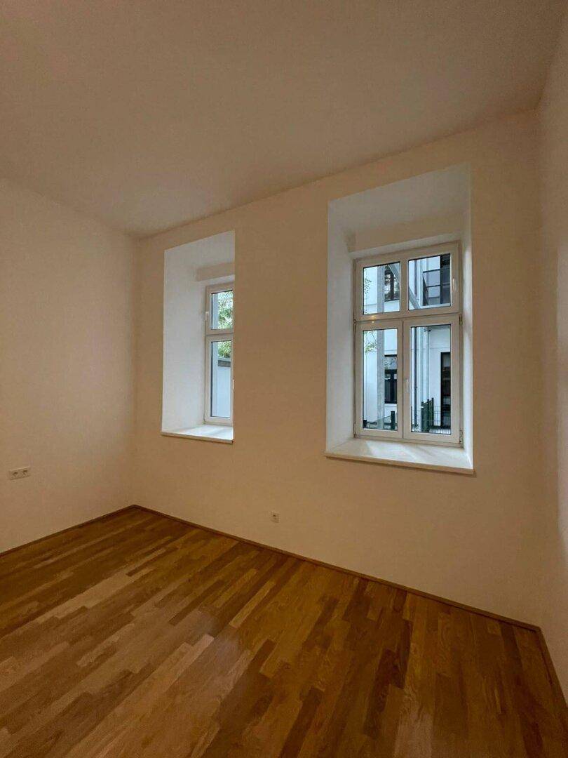 Heller Raum mit Holzboden und zwei Fenstern, die viel Tageslicht hereinlassen.