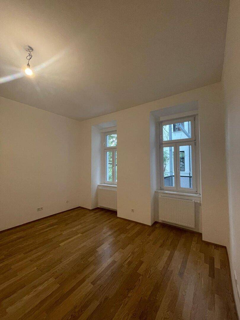 Helles Zimmer mit neuem Parkettboden, weißen Wänden und zwei Fenstern, bereit zum Einzug.