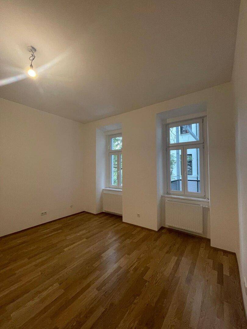 Helles Zimmer mit neuem Parkettboden, weißen Wänden und zwei Fenstern, bereit zum Einzug.