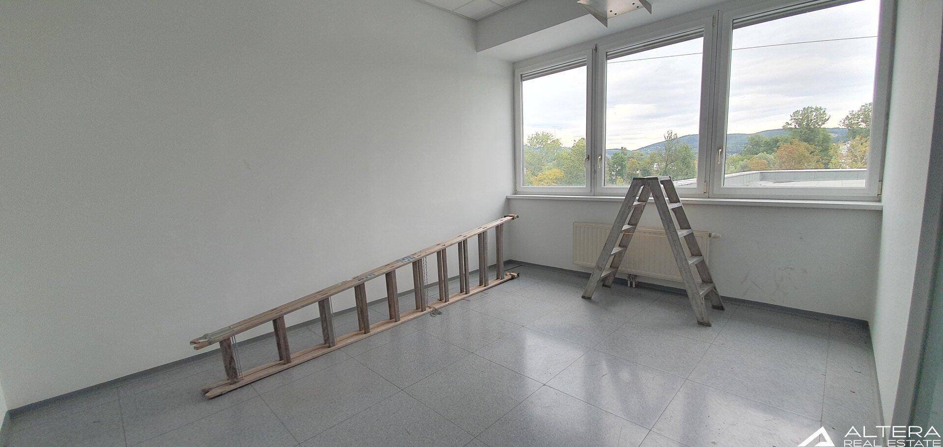 Geräumiger Büroraum mit großen Fenstern und Blick ins Grüne, derzeit mit Leitern für Arbeiten.