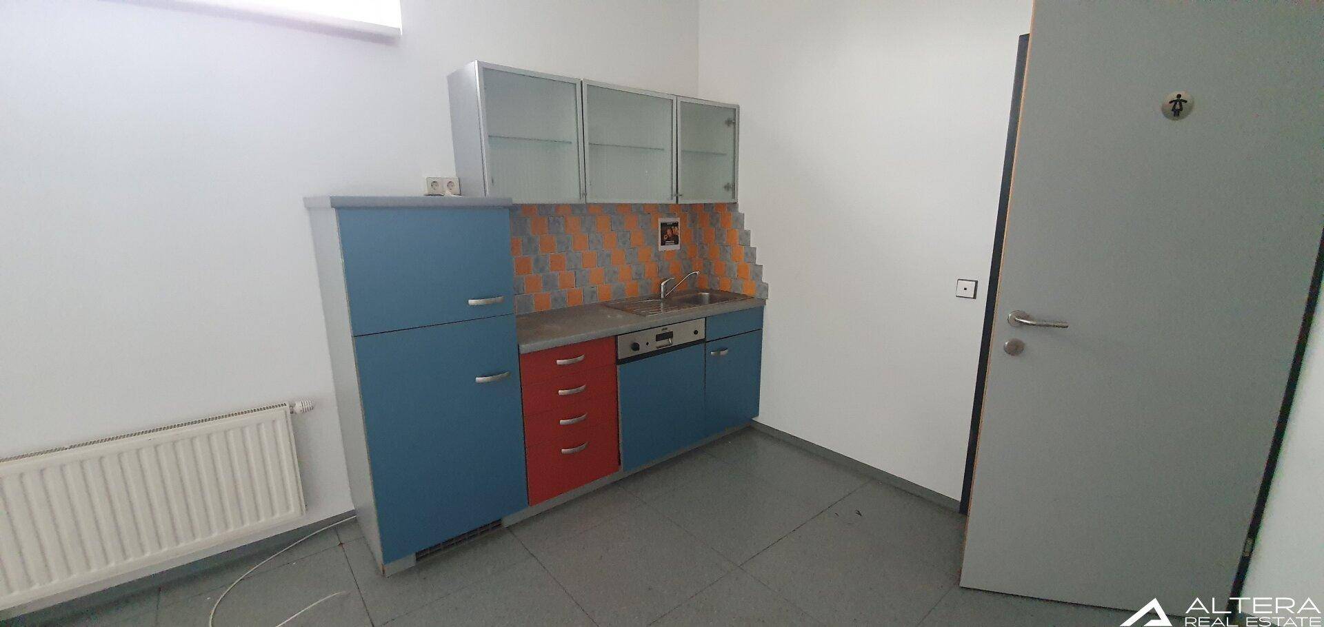 Funktionale Küchenzeile mit blauen und roten Schränken, Spüle und Fliesenrückwand in einem Büroraum.