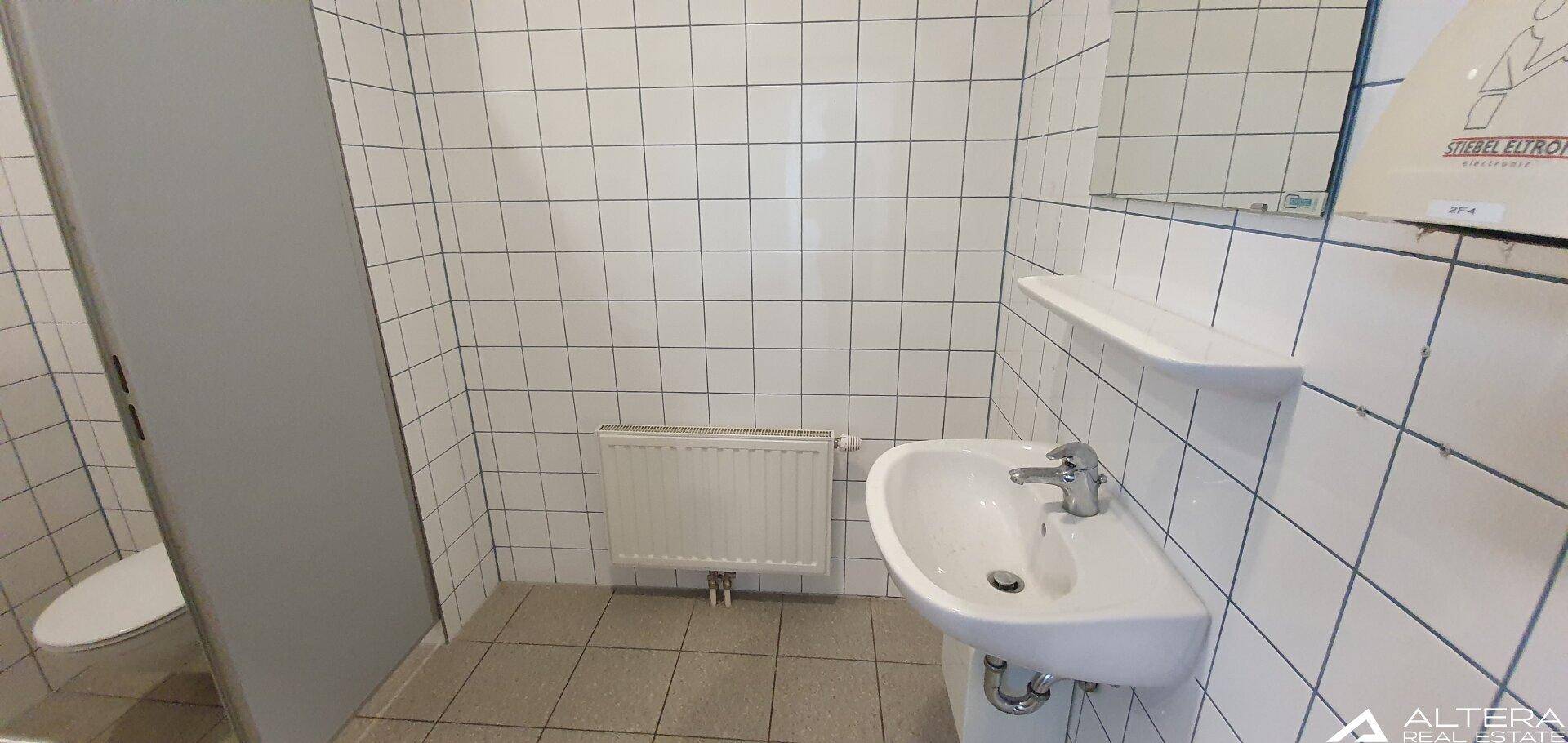 Einfache Toilette mit weißen Fliesen an Wänden und Boden, Waschbecken und Heizkörper.