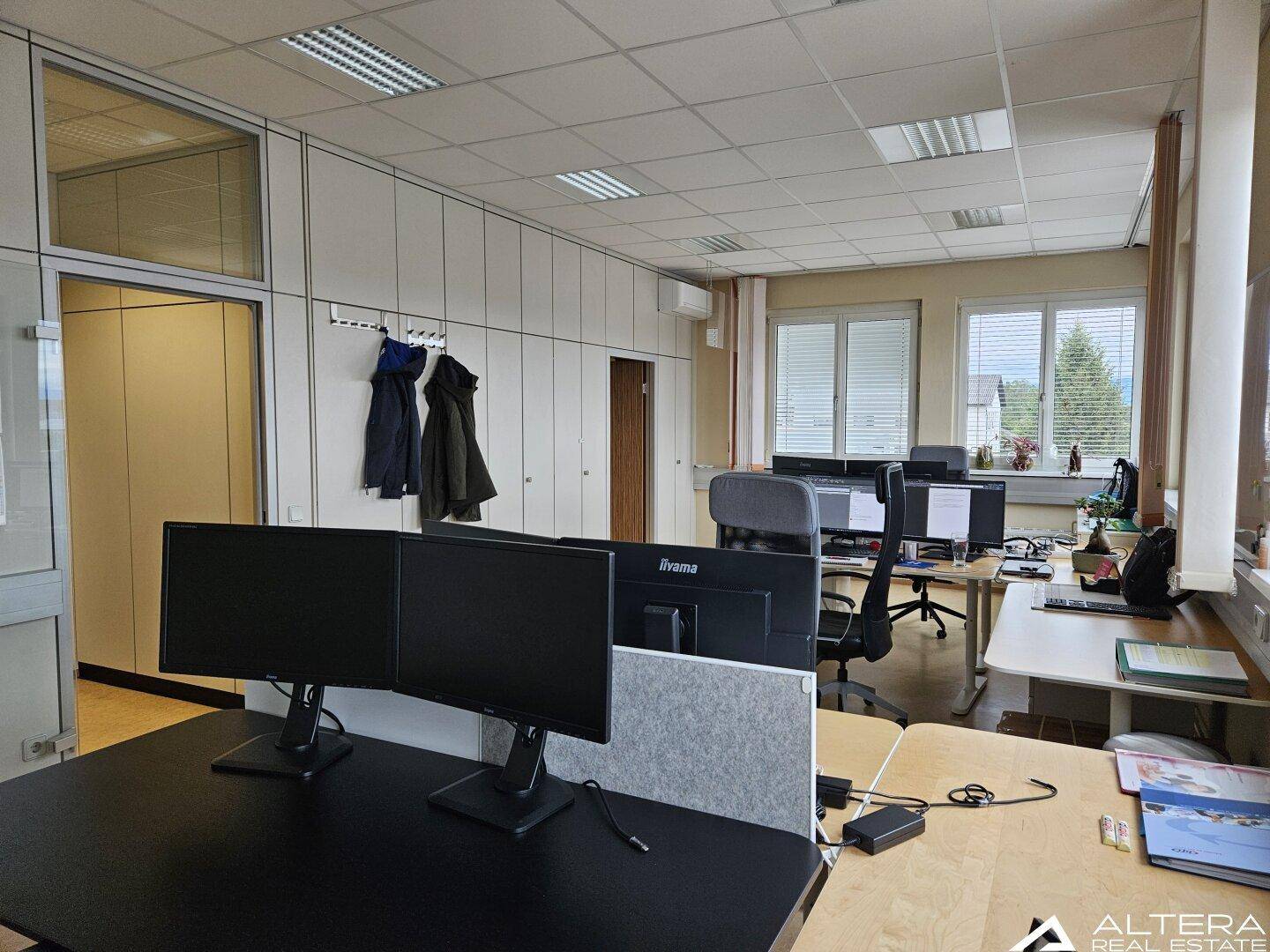 Büro mit mehreren Arbeitsplätzen, großen Fenstern und einer Wand mit Garderobenhaken.