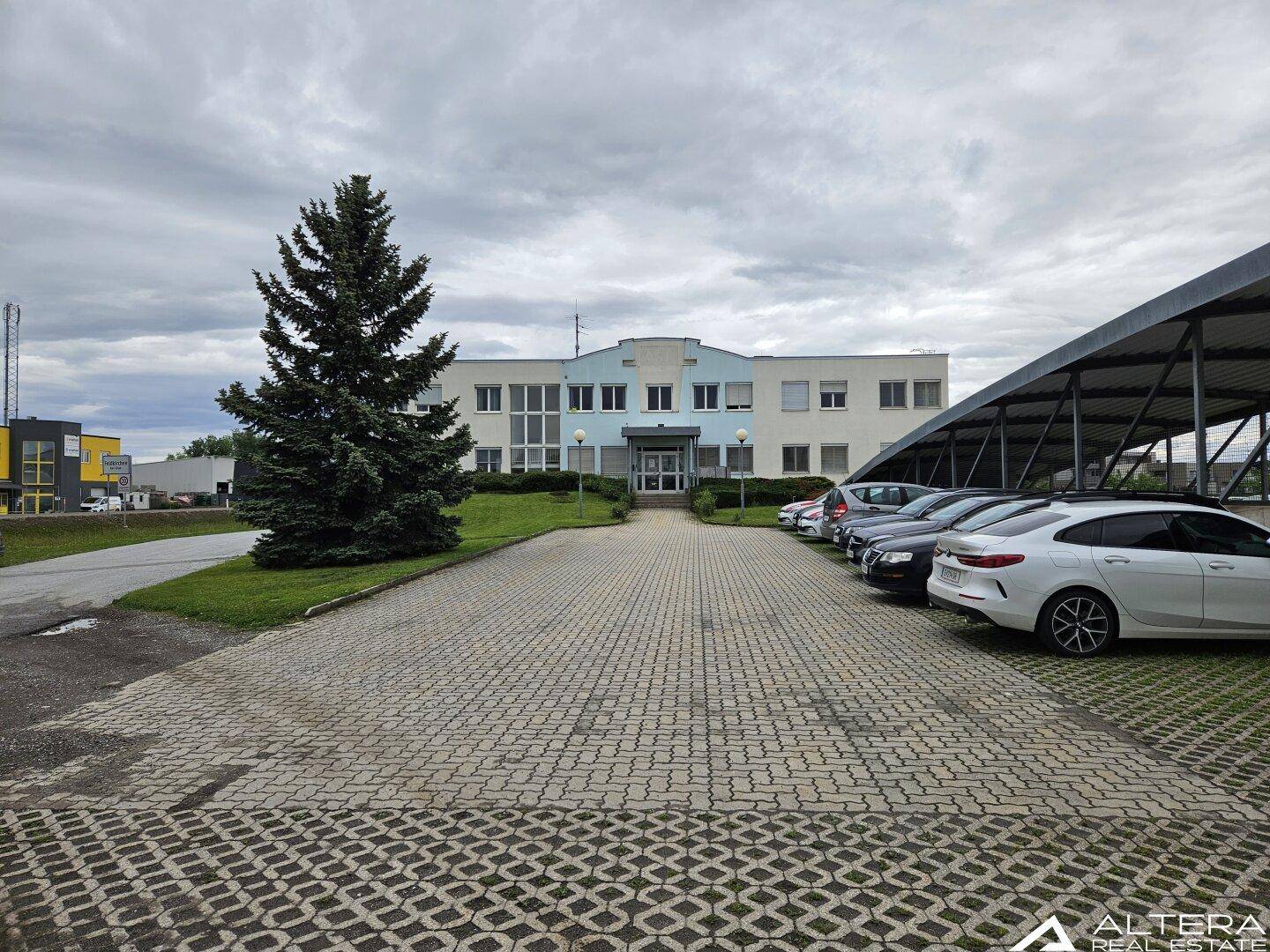 Außenansicht eines Bürogebäudes mit Parkplätzen und einem Carport unter bewölktem Himmel.