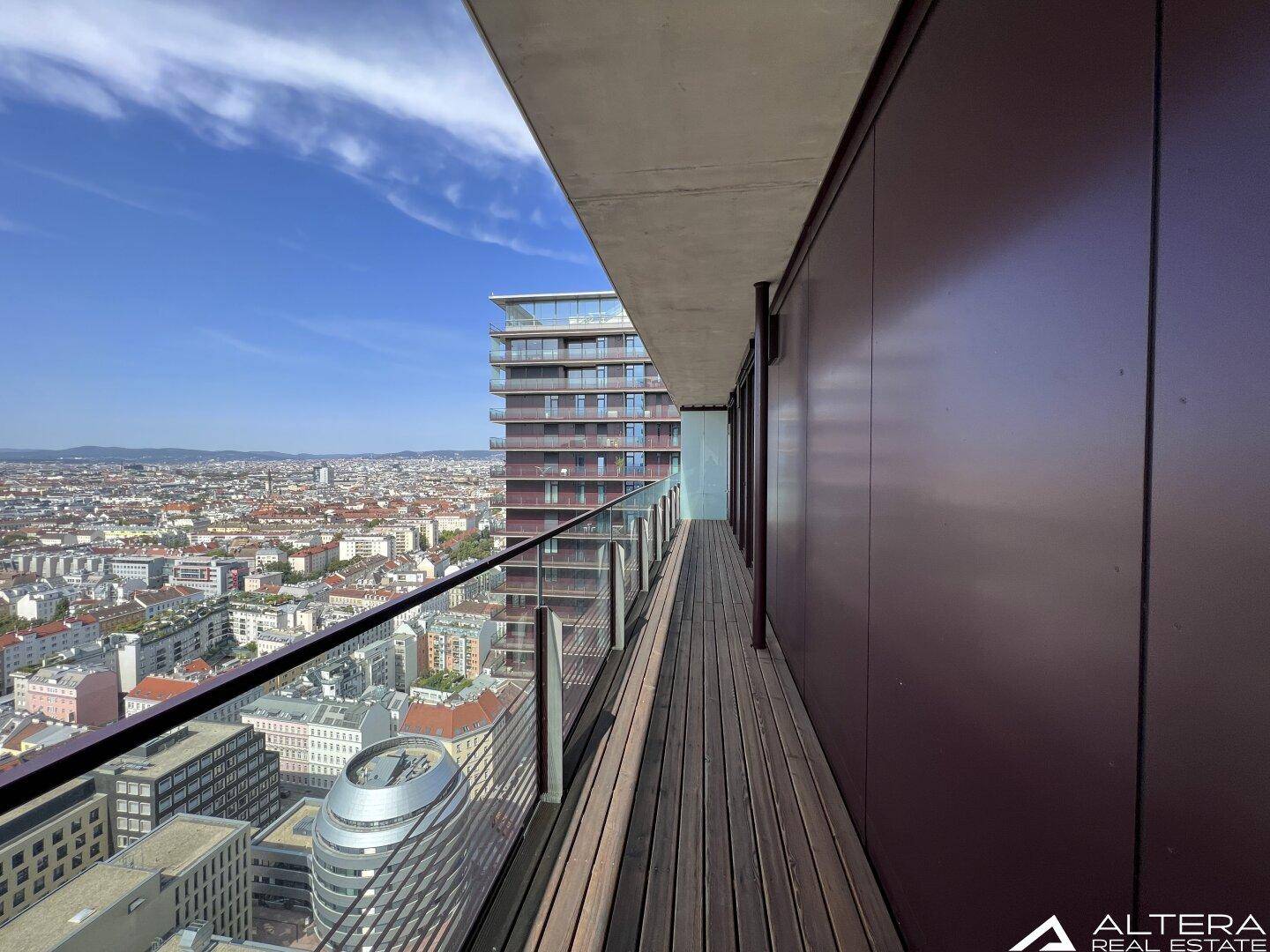 Langer Balkon mit Holzdeck und Glasgeländer, bietet weite Aussicht über die Stadt.