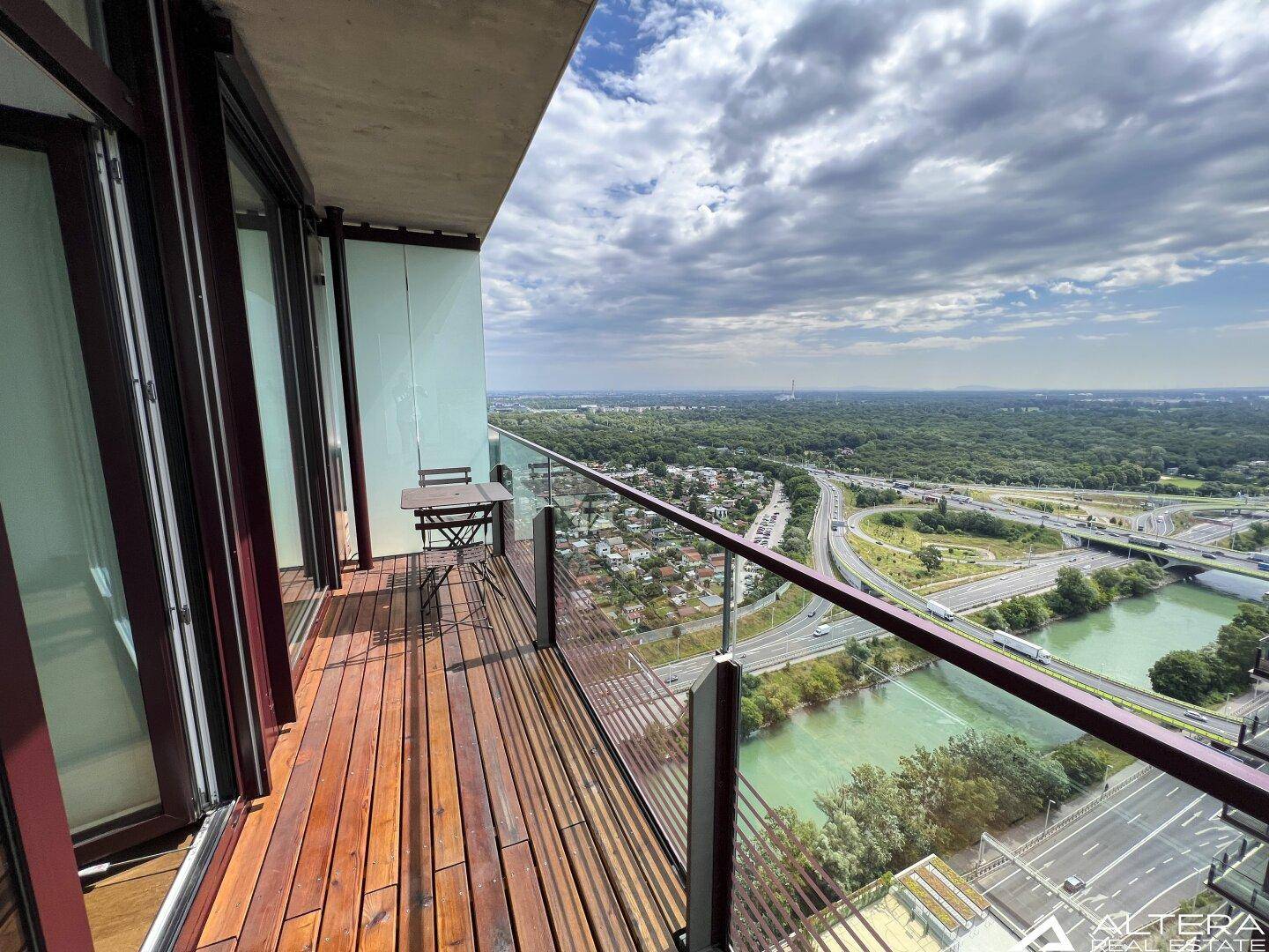 Langer Balkon mit Holzdeck, Glasgeländer und weitem Blick über Stadt und Fluss.
