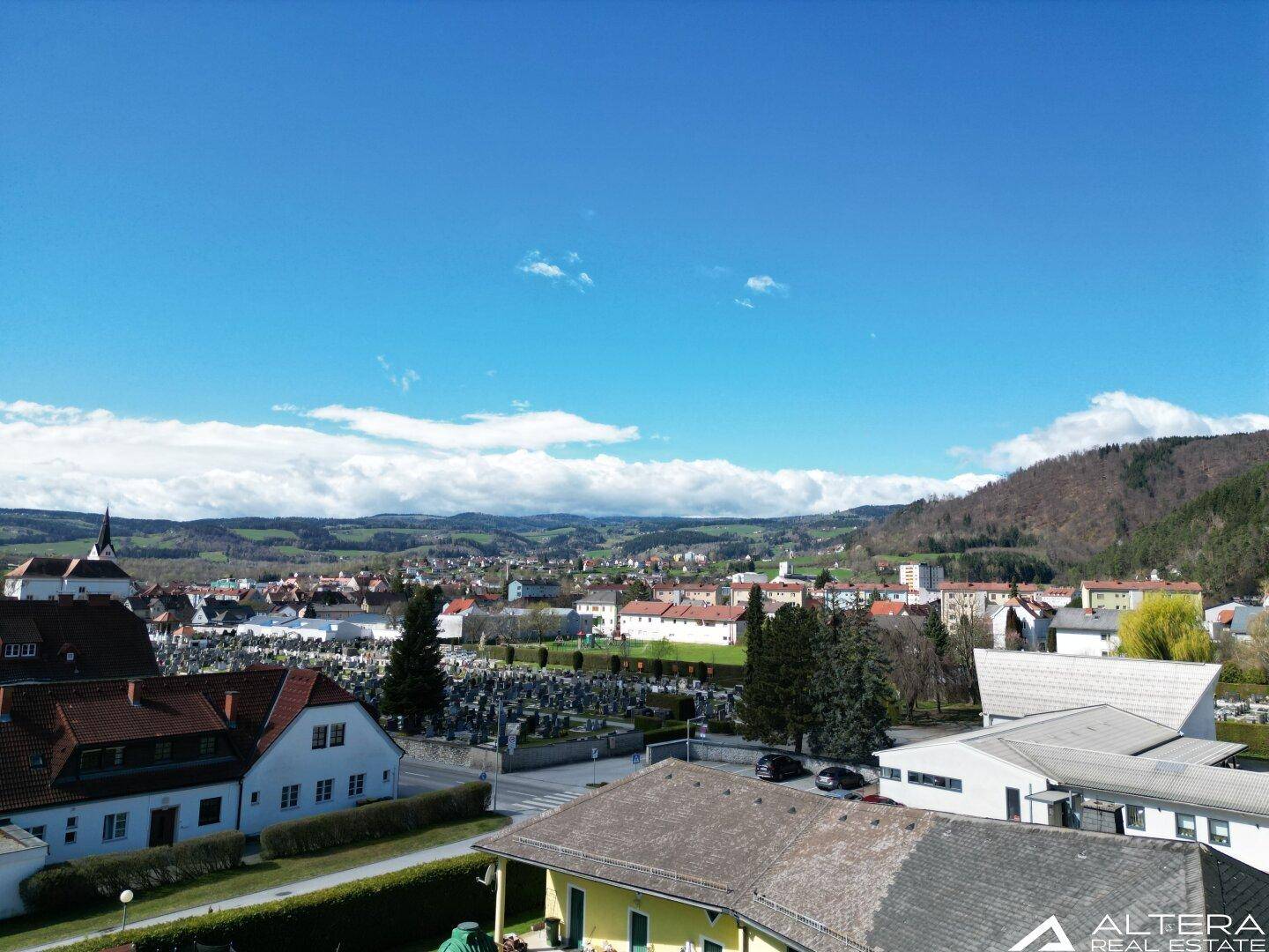 Weitläufiger Blick über eine ländliche Stadt mit Friedhof und hügeliger Landschaft im Hintergrund unter blauem Himmel.