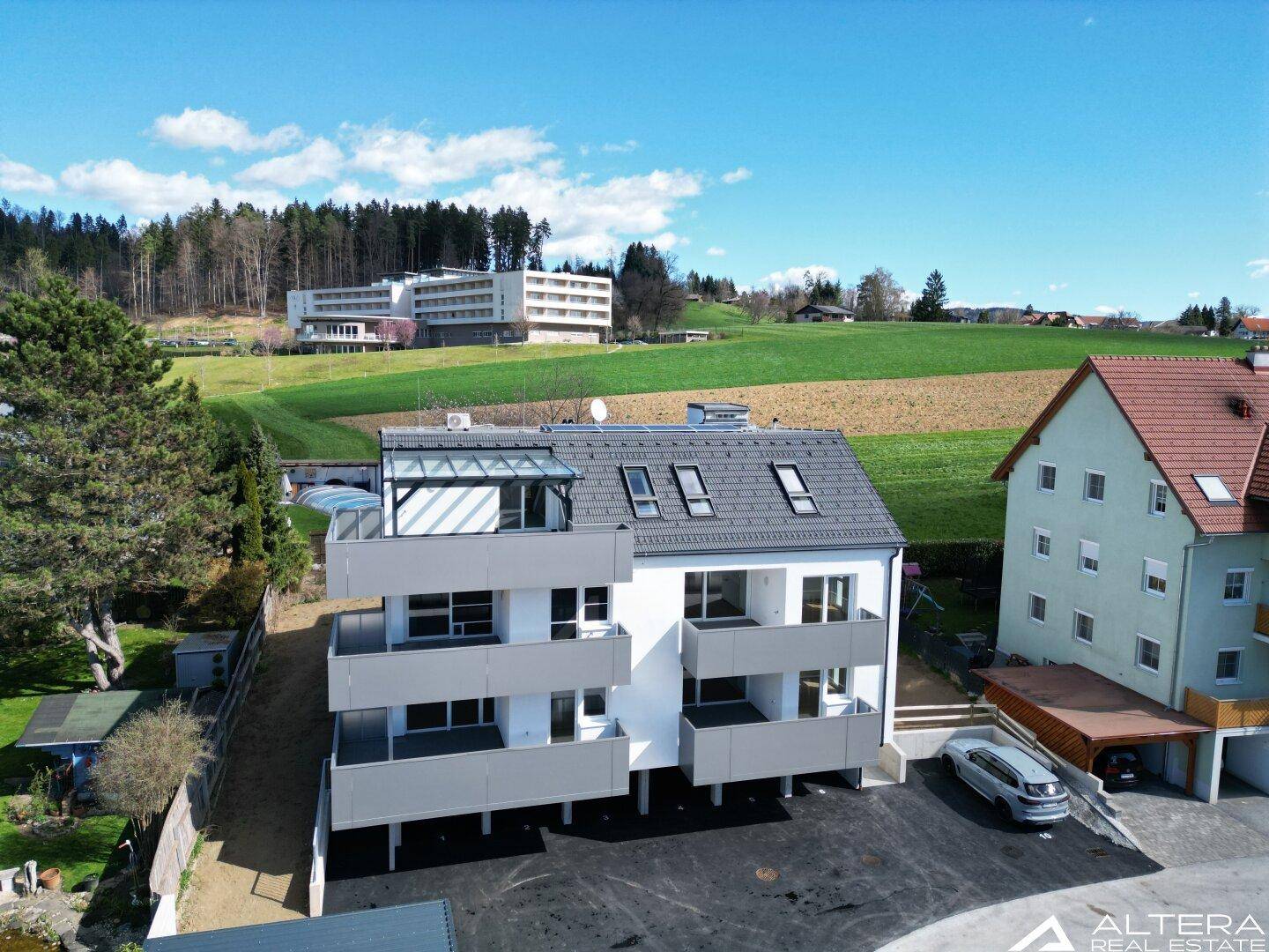 Luftaufnahme eines modernen Apartmentgebäudes mit Balkonen und Dachterrasse in grüner Umgebung.