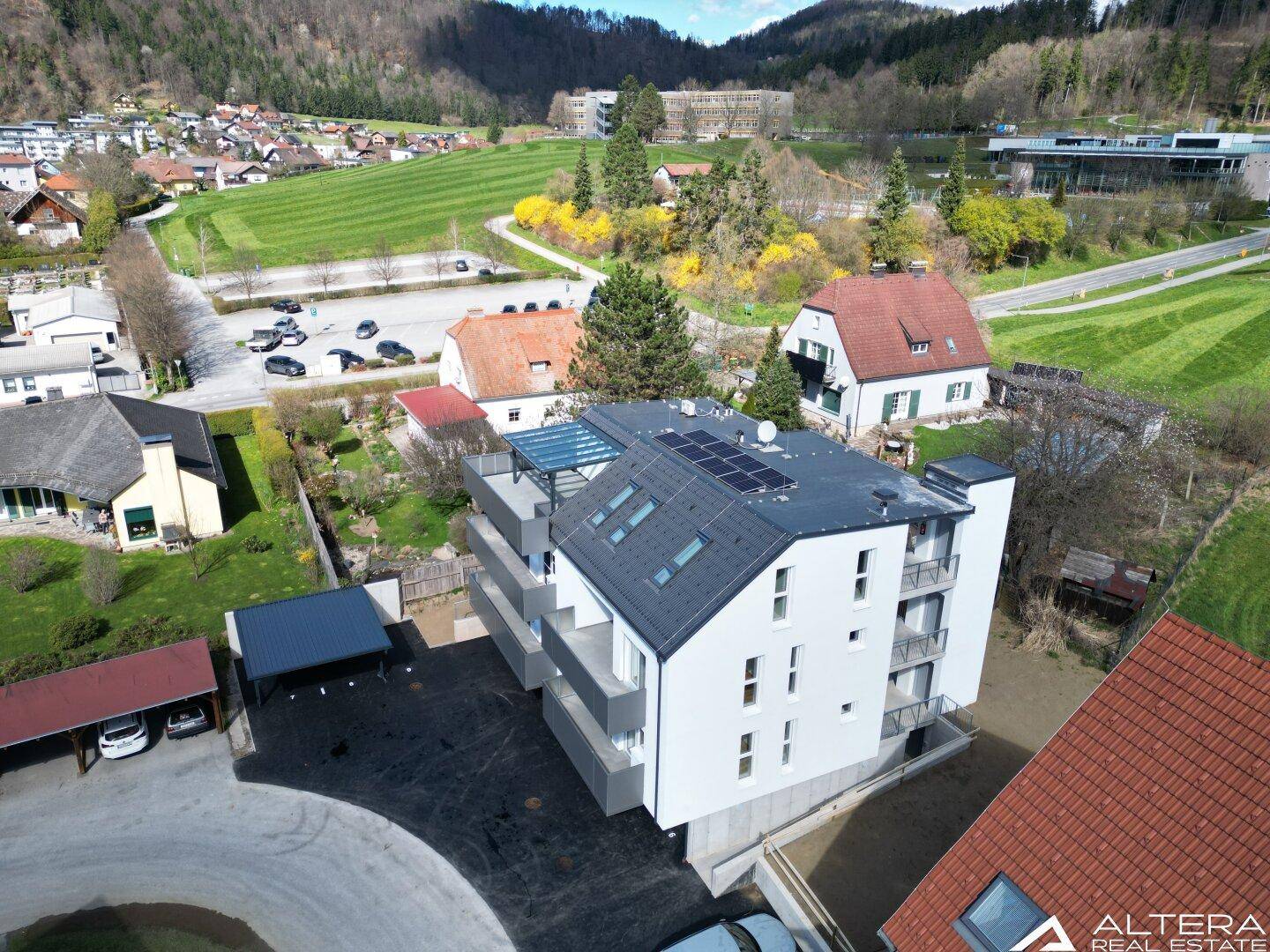 Luftaufnahme eines modernen Neubaus mit Solaranlage und umliegender Wohnbebauung in grüner Umgebung.