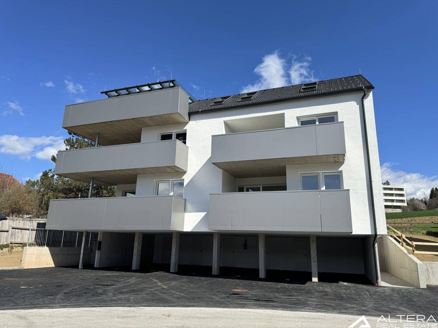 Moderne Fassade eines Neubaus mit mehreren Balkonen und Zugang zur Tiefgarage unter strahlend blauem Himmel.
