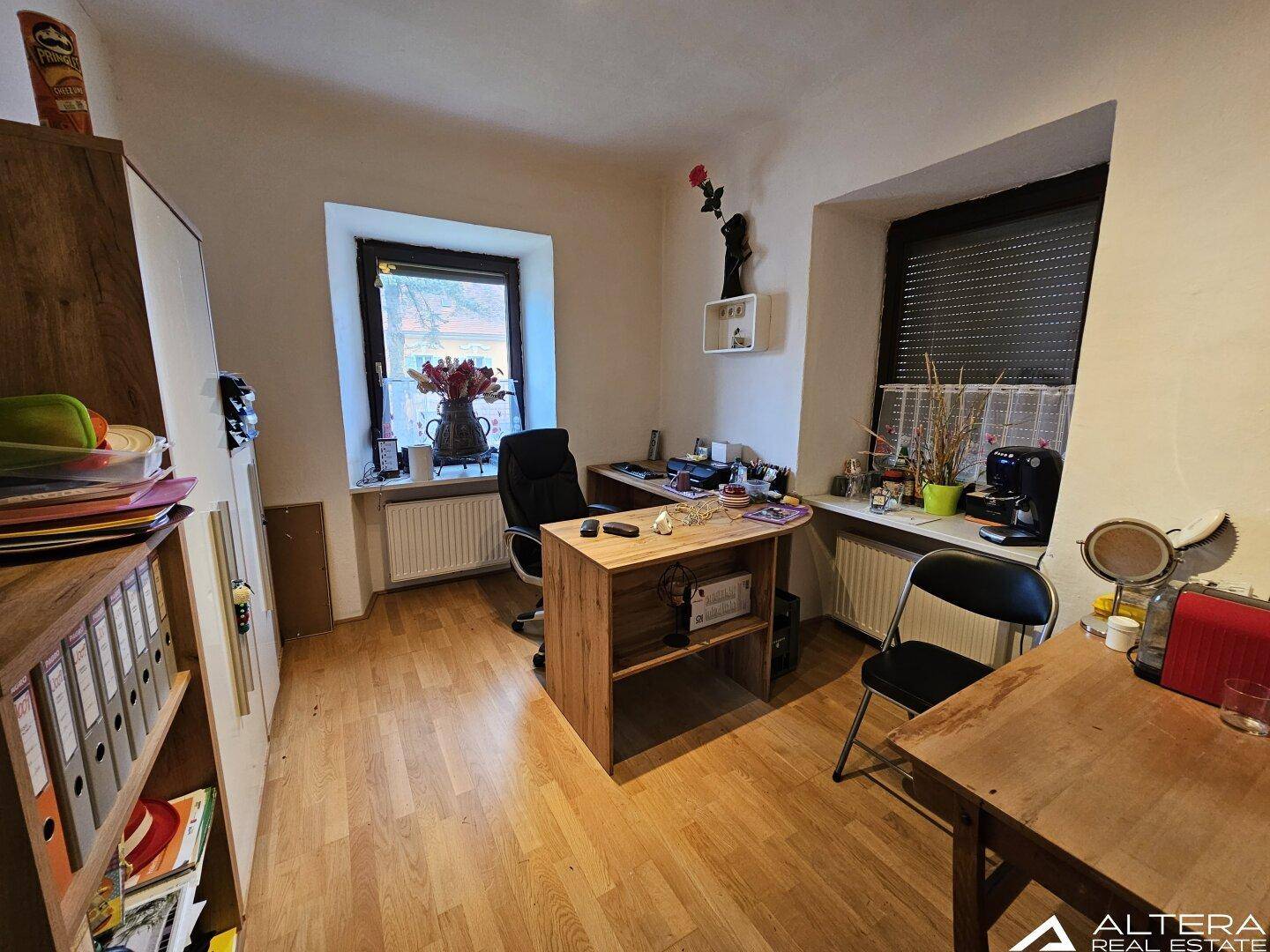 Helles Arbeitszimmer mit Holzparkett, zwei Fenstern und einem Schreibtischbereich.
