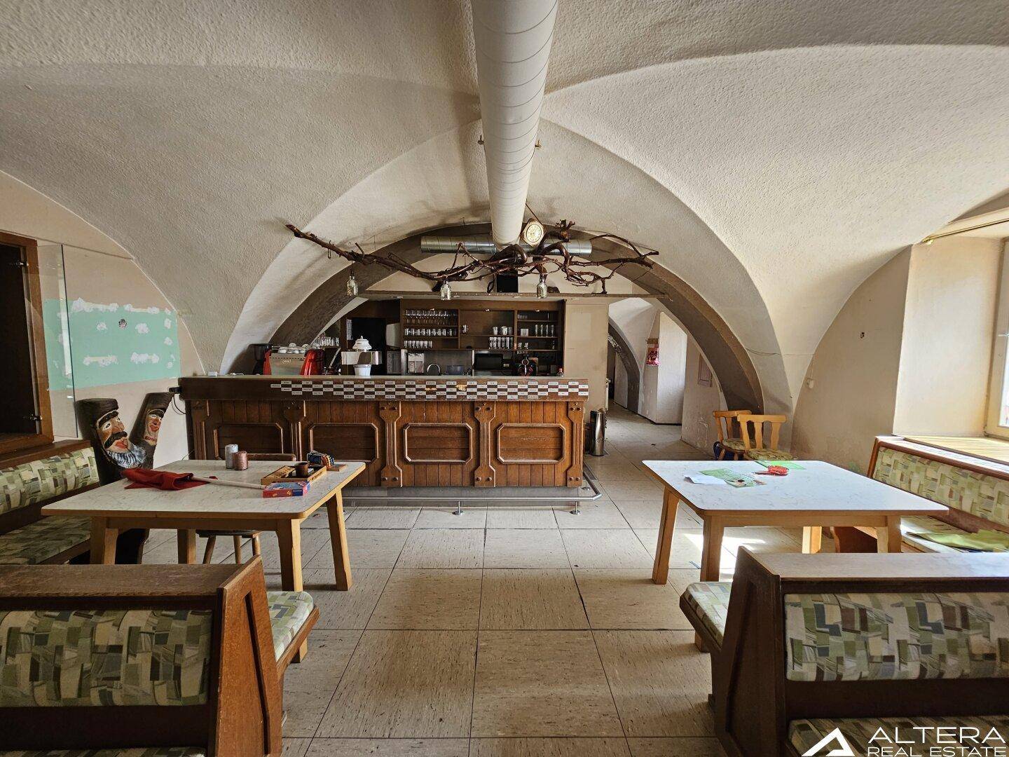 Innenraum mit Gewölbedecke, Holztischen und einer Bar im Hintergrund, die eine rustikale Atmosphäre schafft.