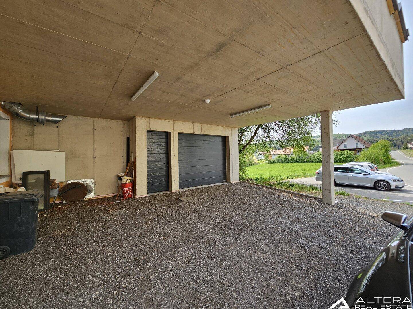 Überdachter Bereich mit Garagentoren und Parkmöglichkeiten, umgeben von Grünflächen.