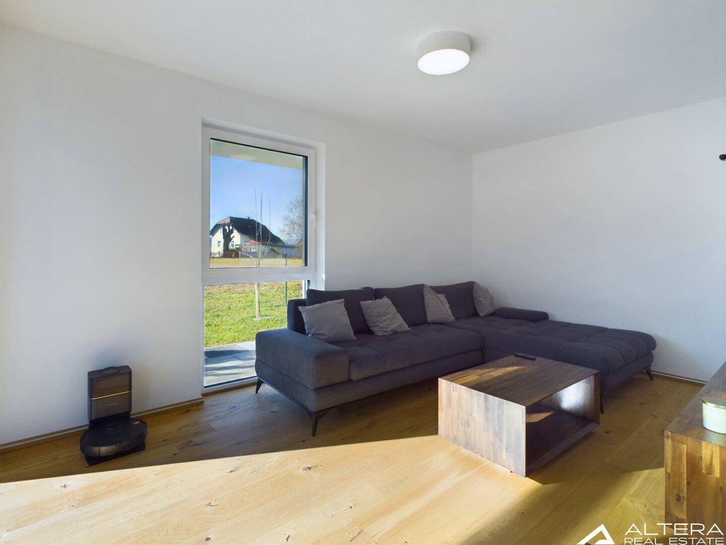 Gemütliches Wohnzimmer mit grauem Ecksofa, Holzboden und einem großen Fenster mit Blick ins Grüne.