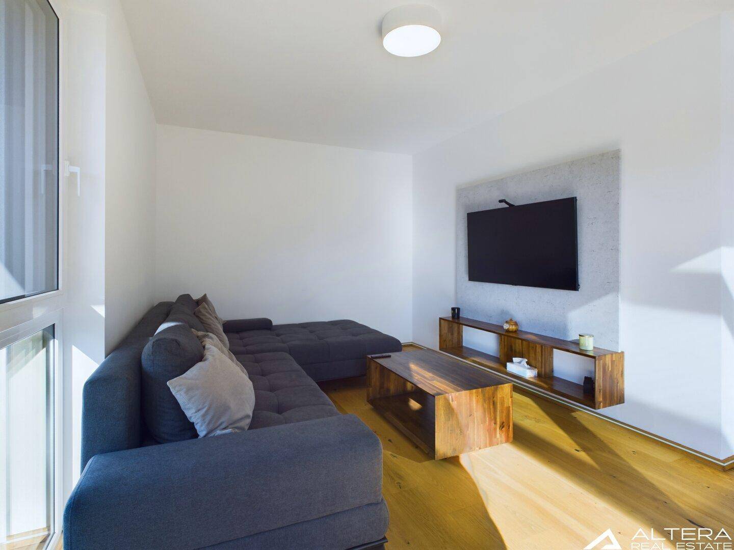 Modern eingerichtetes Wohnzimmer mit großem Sofa, Holzboden und einem an der Wand montierten Fernseher.