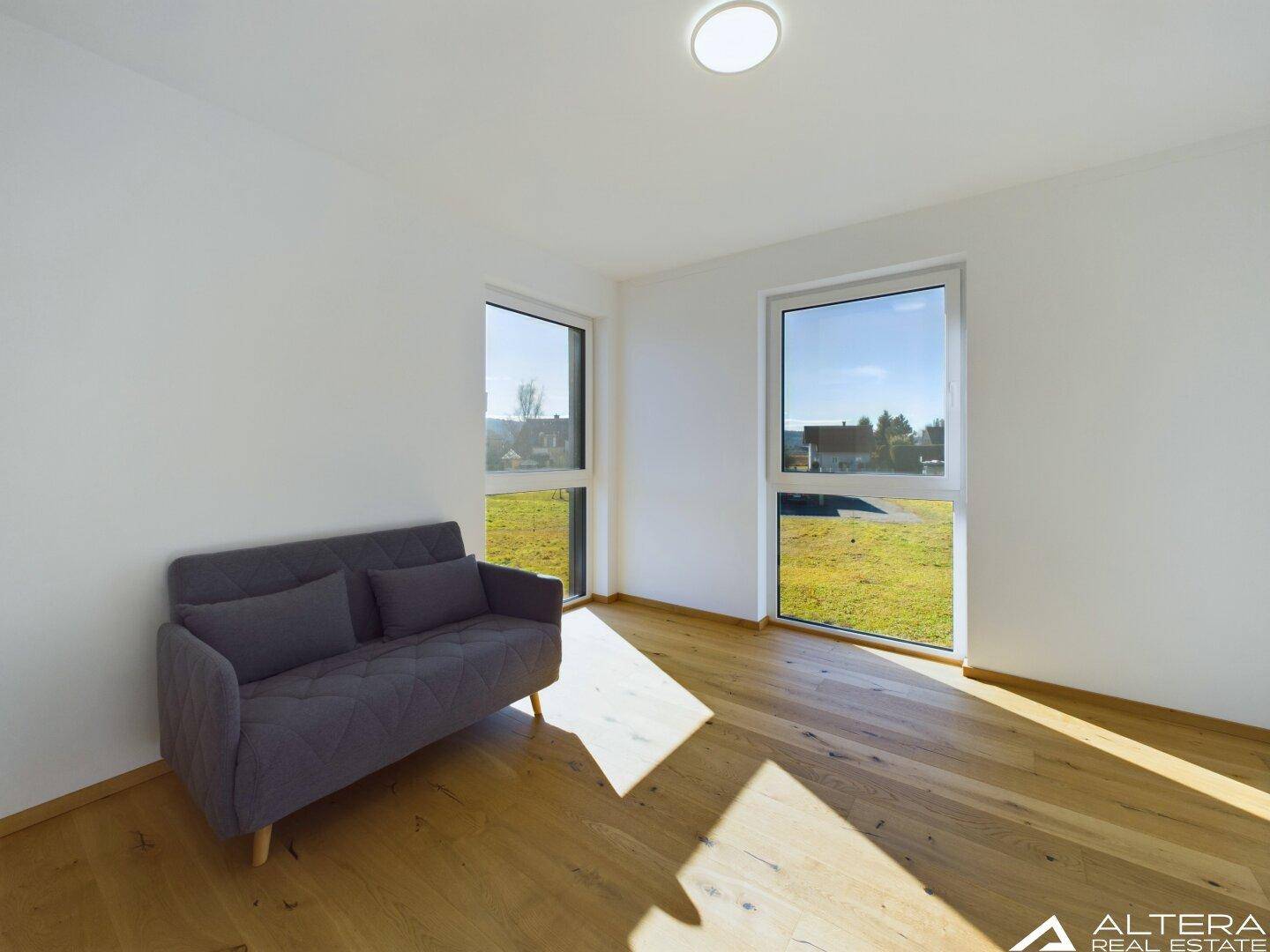 Helles Schlafzimmer mit Holzboden, kleinem Sofa und großen Fenstern mit Blick ins Grüne.