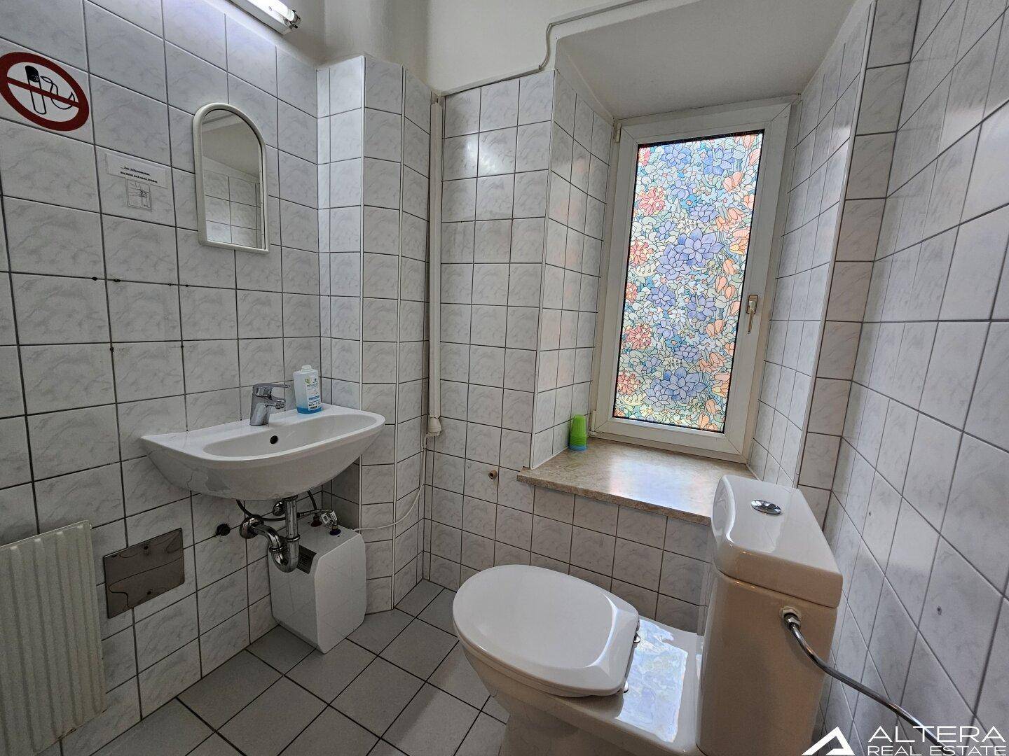 Gefliestes Badezimmer mit Waschbecken, Toilette und einem Fenster mit farbigem Glasdesign.