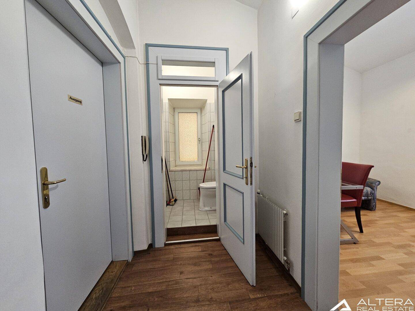 Flur mit dunklem Holzboden, der zu einem Badezimmer mit Fliesen und Fenster führt.