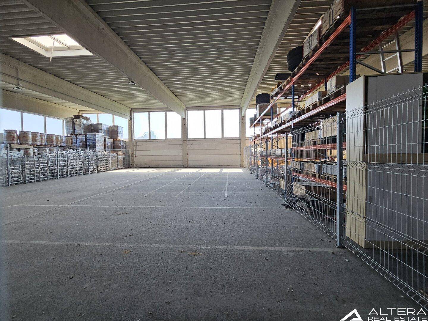 Weitläufige Lagerhalle mit Betonboden, hohen Decken und großen Fenstern, die viel Tageslicht hereinlassen.