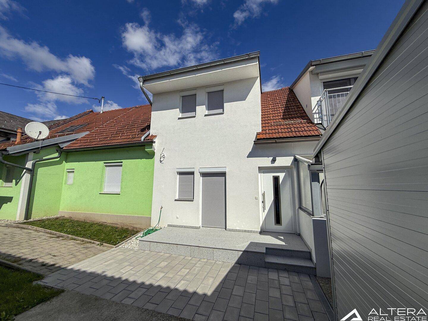 Vorderansicht des Hauses mit Eingangstür, Rollläden und gepflastertem Vorplatz an einem sonnigen Tag.