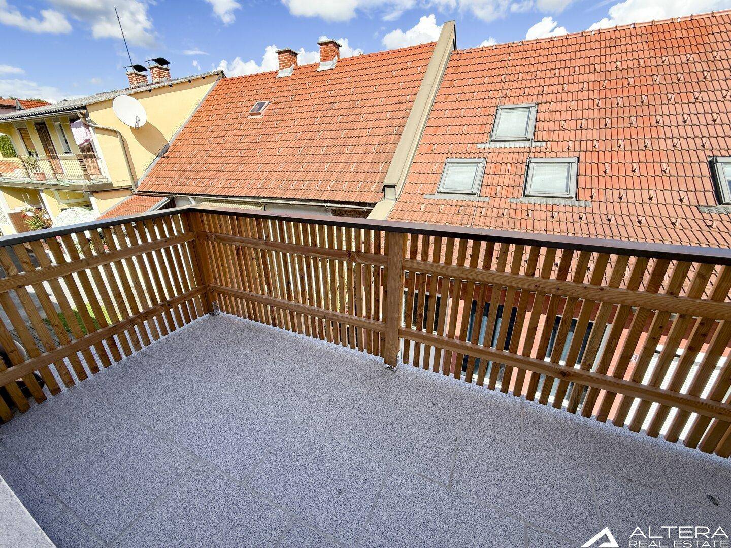 Geräumiger Balkon mit Holzzaun und Blick auf die umliegenden Dächer und den Himmel.