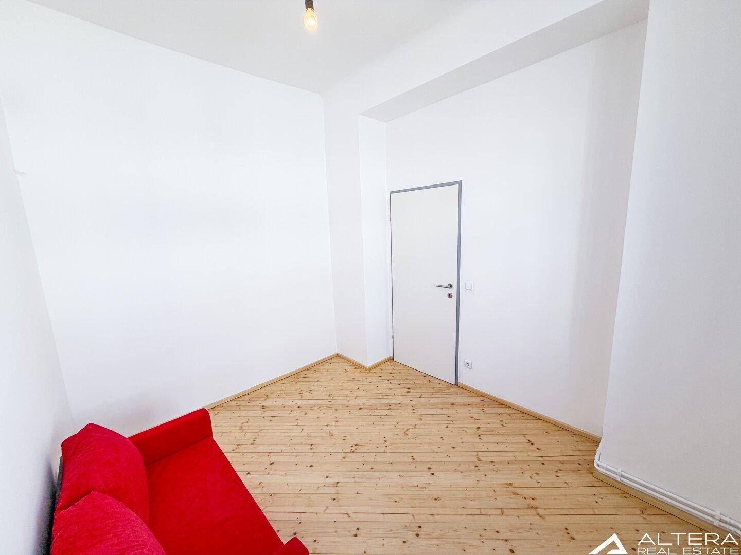 Gemütlicher Raum mit rotem Sofa, Holzdielenboden und einer modernen weißen Tür.