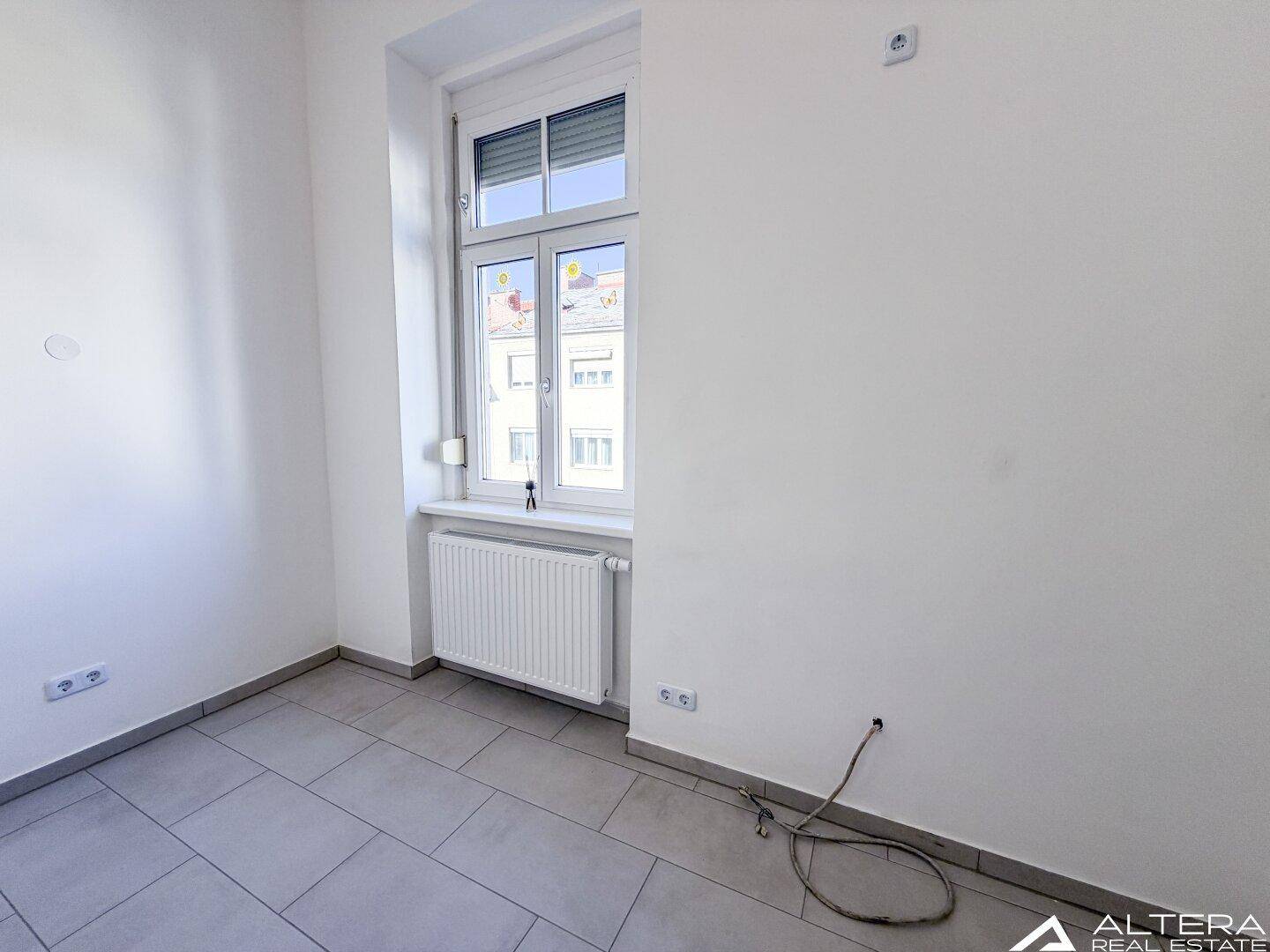 Heller Raum mit grauen Fliesen und einem Fenster, geeignet als kleines Büro oder Abstellraum.