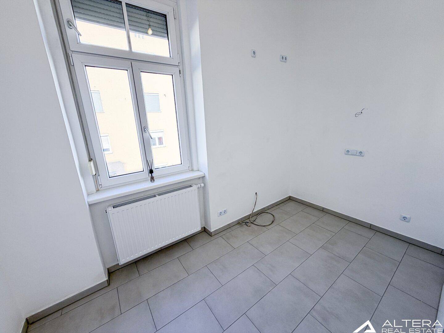 Detailansicht eines Raumes mit grauen Fliesenboden und einem Fenster, zeigt Steckdosen und Kabel.