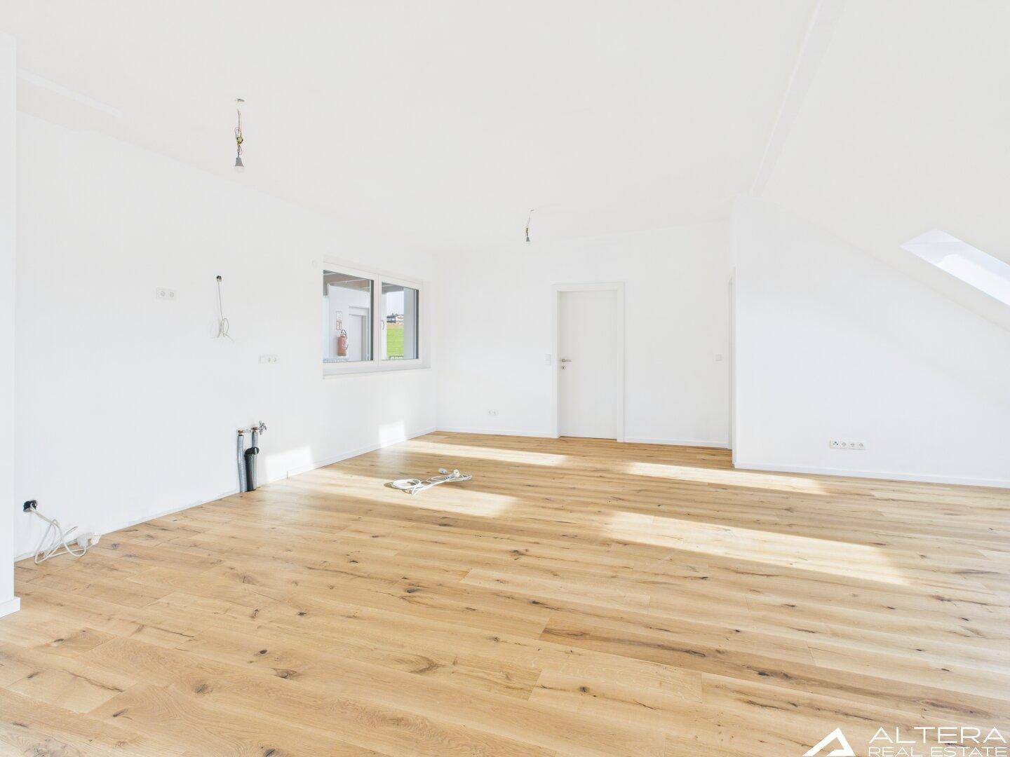 Freundliches Zimmer mit Holzboden, weiß gestrichenen Wänden und einem Fenster mit Blick ins Grüne.