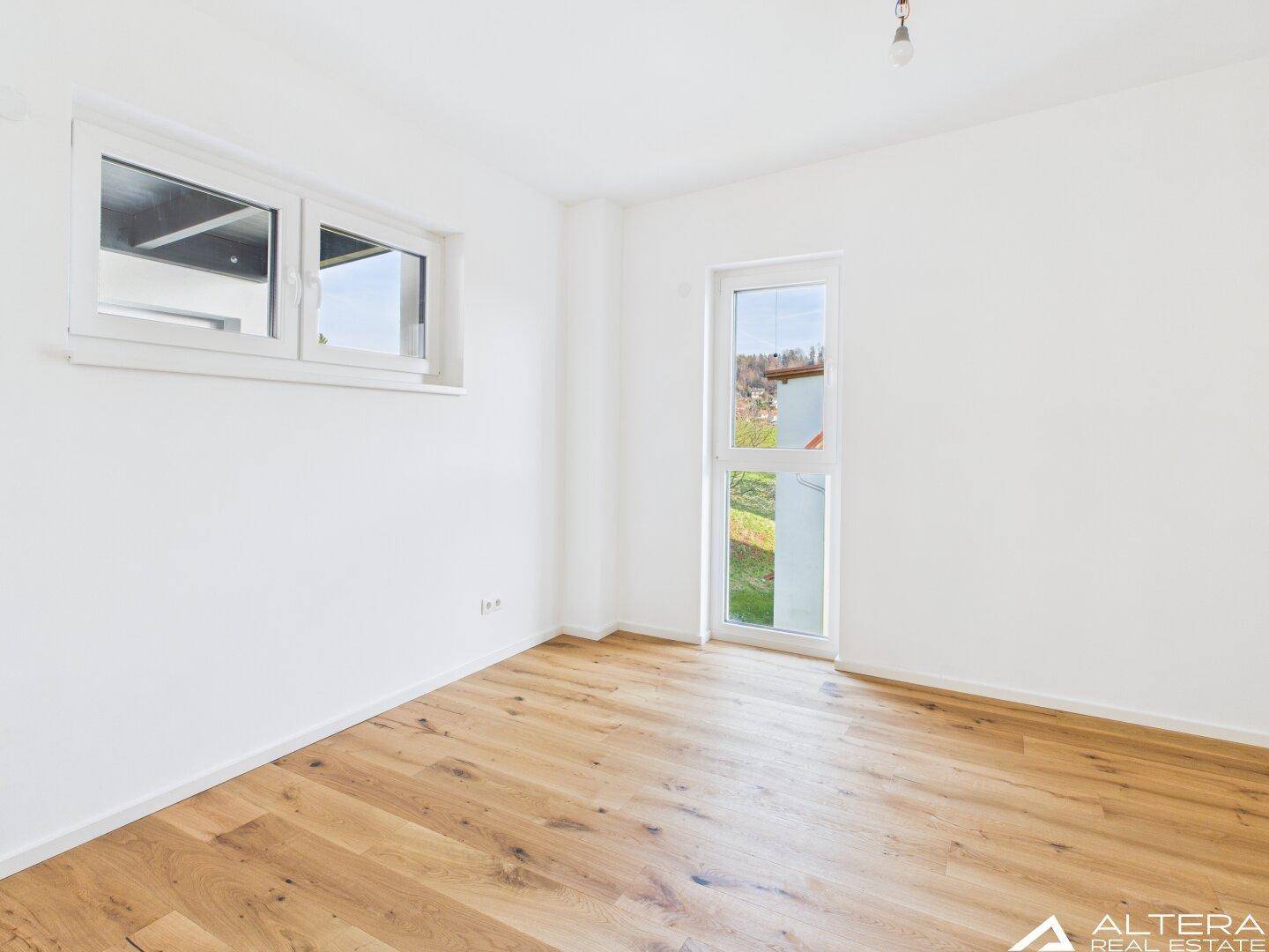 Helles Zimmer mit Holzboden und zwei Fenstern, die viel Tageslicht hereinlassen.