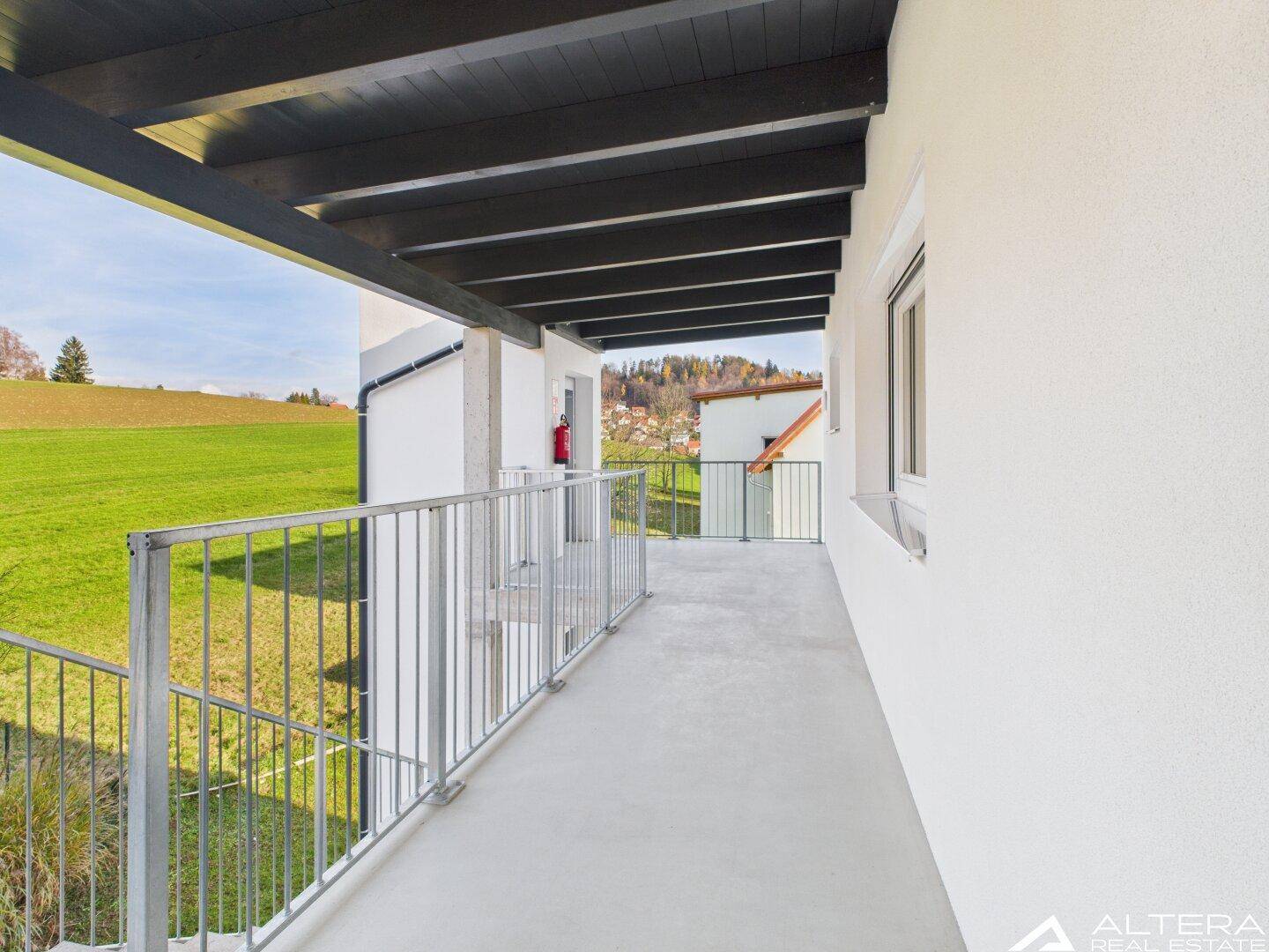 Langer, überdachter Balkon mit Metallgeländer und Blick auf grüne Felder und die Umgebung.