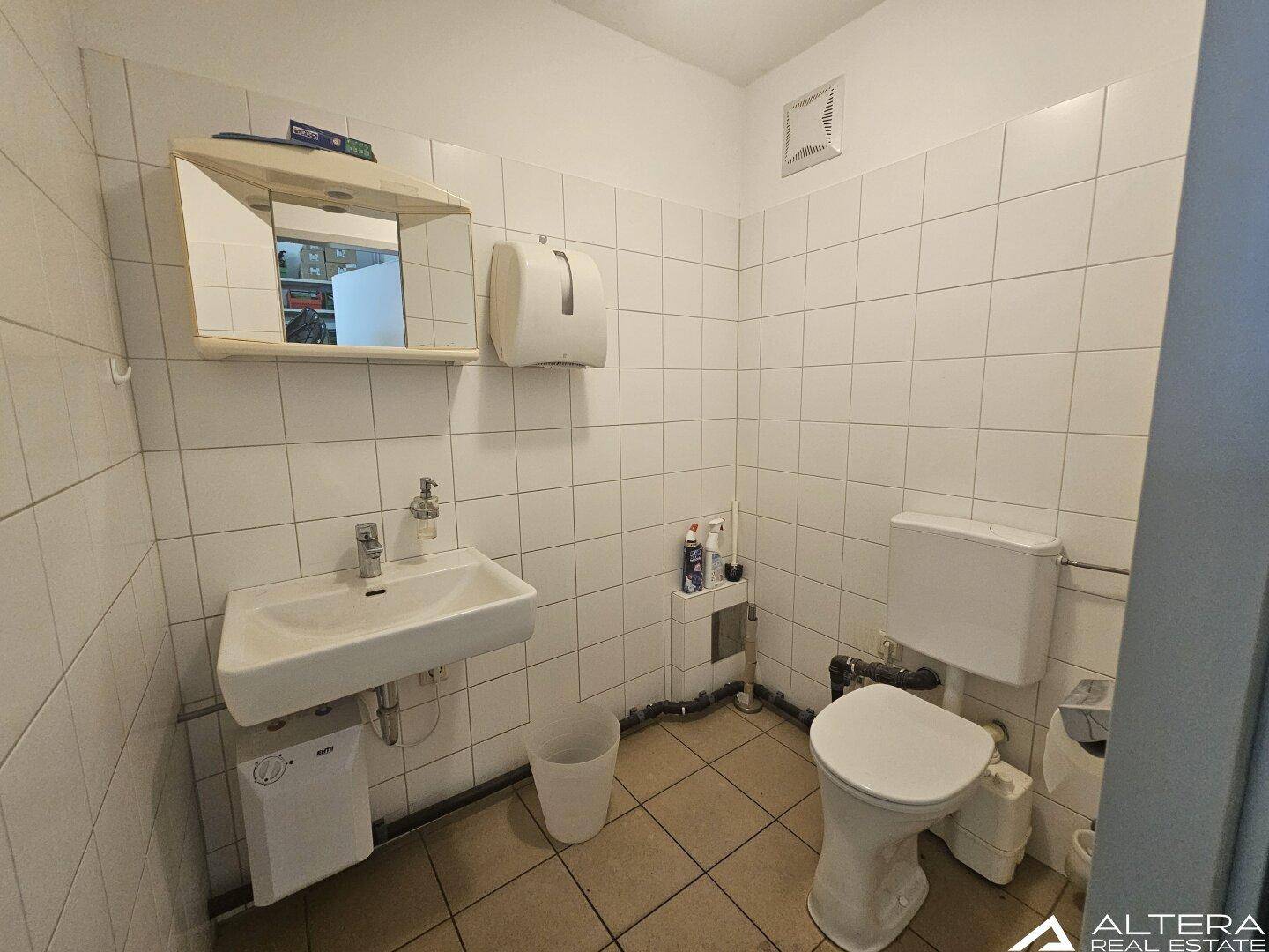 Einfache Toilette mit weißen Fliesen, Waschbecken und Spülkasten, ausgestattet mit einem Wandspiegel.