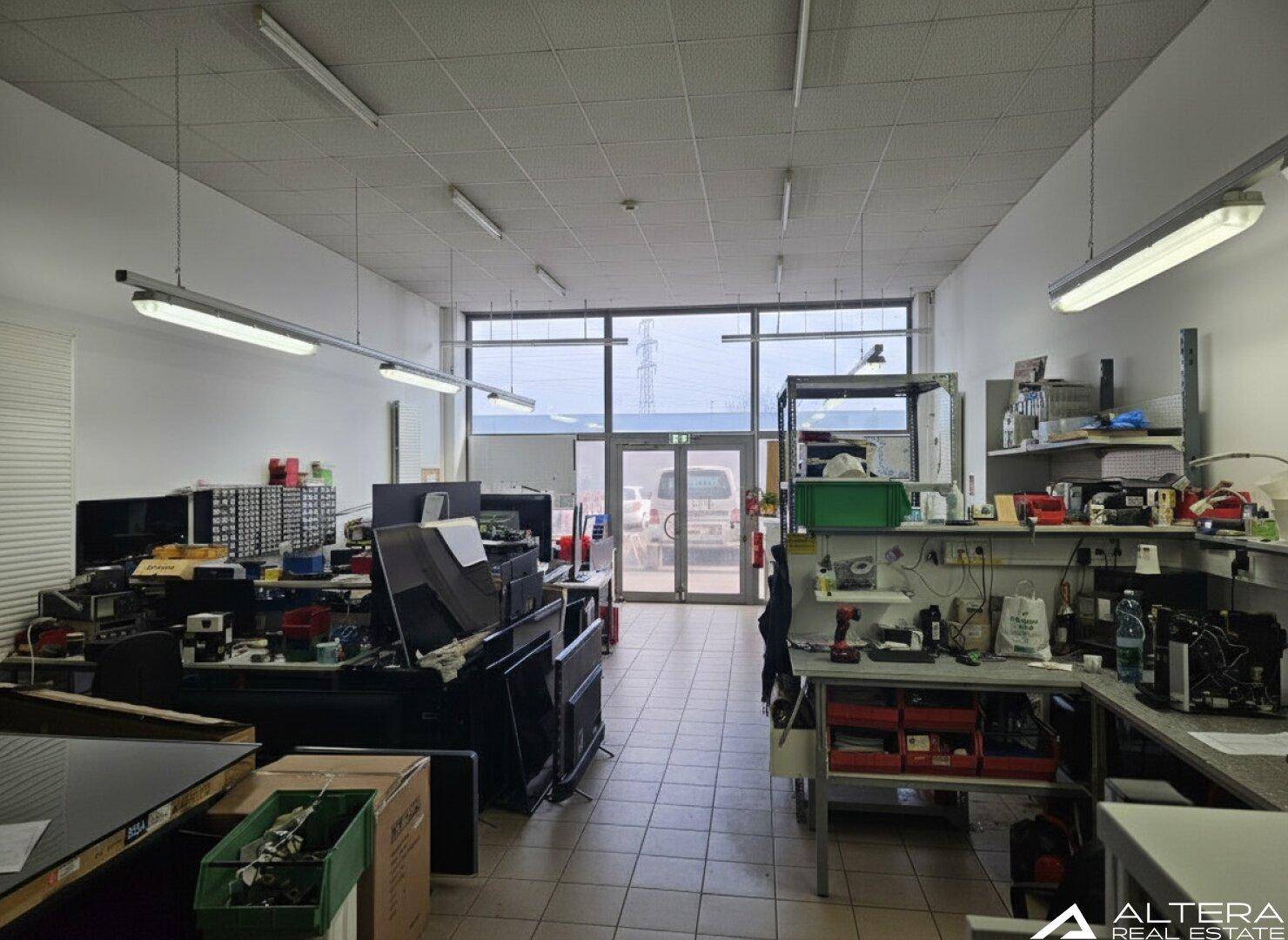 Heller Werkstattbereich mit großen Fenstern am Eingang, ausgestattet mit Arbeitsplätzen und Regalen.