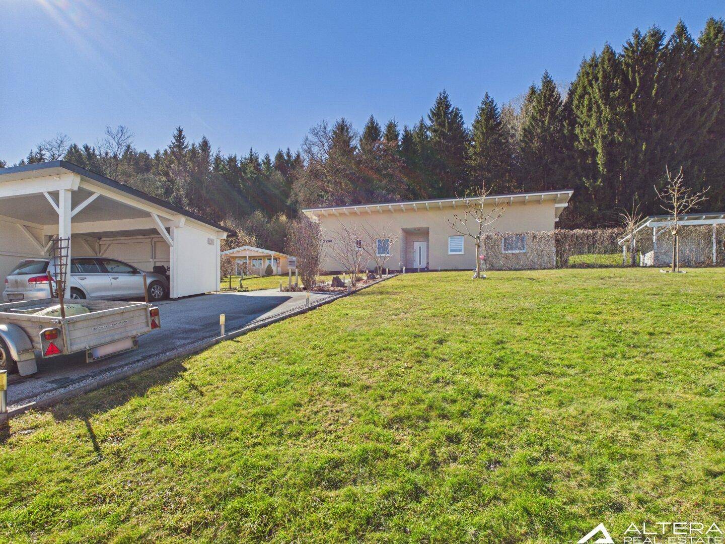Gesamtansicht des Hauses mit Carport, großem Rasen und Wald im Hintergrund.