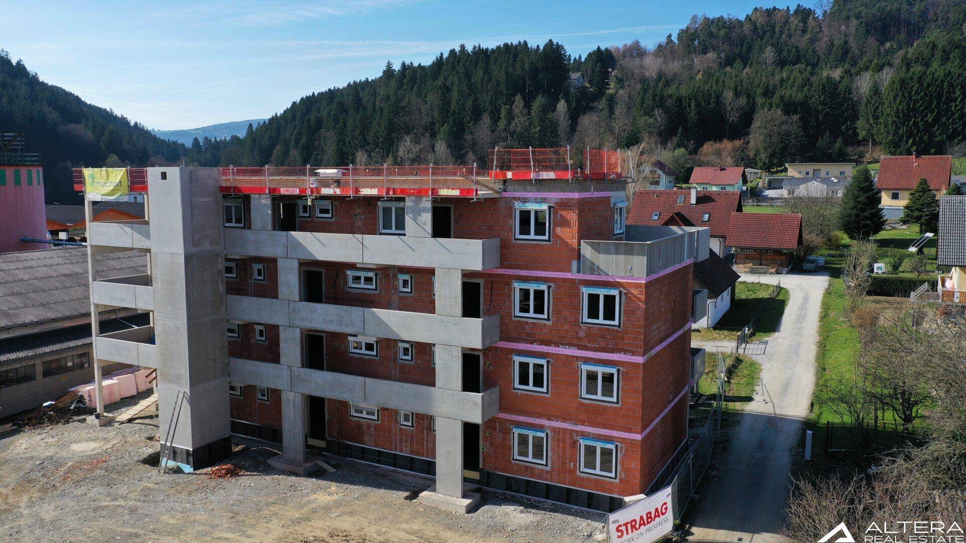 Neubau im Rohbau mit Fenstern und Blick auf bewaldete Hügel und umliegende Häuser.