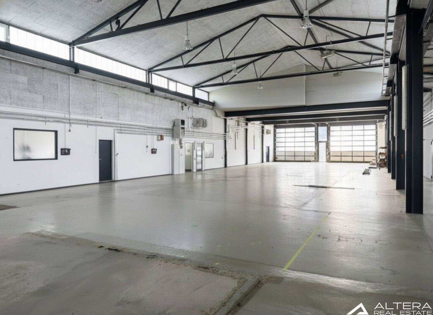 Große, leere Industriehalle mit hohen Decken, Betonboden und viel natürlichem Licht durch Oberlichter.