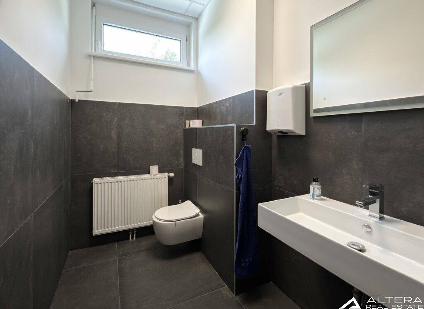 Modernes Gäste-WC mit dunklen Fliesen, wandhängendem WC und Fenster für Tageslicht.