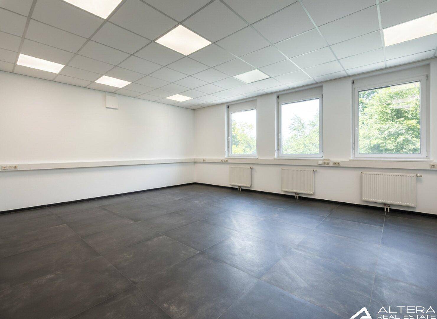 Heller Büroraum mit drei großen Fenstern, dunklem Fliesenboden und weißer Decke.