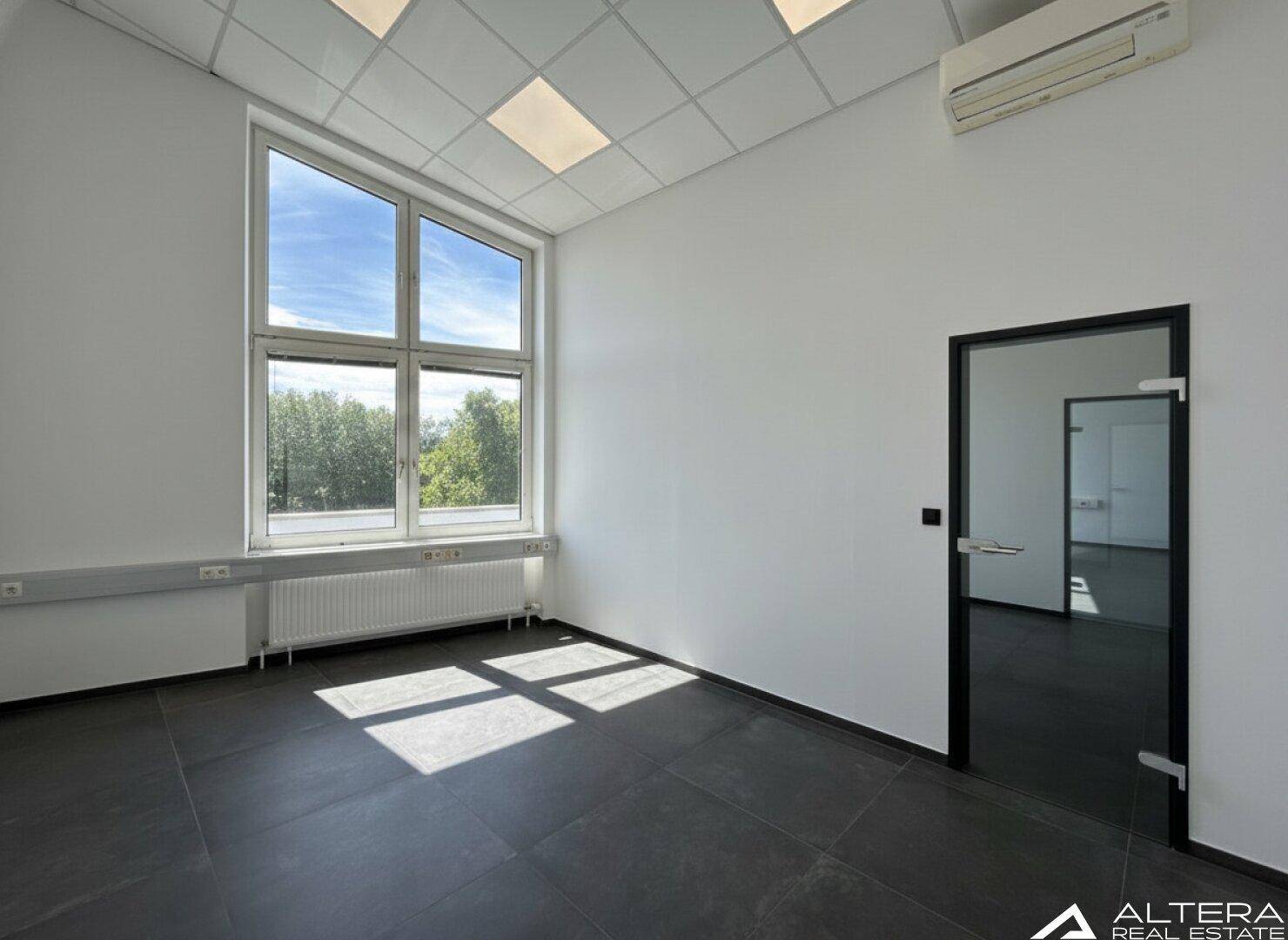 Geräumiger Büroraum mit hohen Fenstern, dunklem Fliesenboden und viel Tageslicht.
