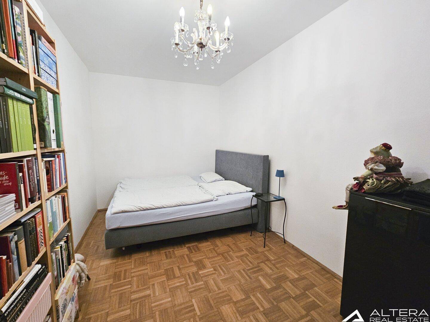 Schlafzimmer mit Parkettboden, einem Bett und einem großen Bücherregal.