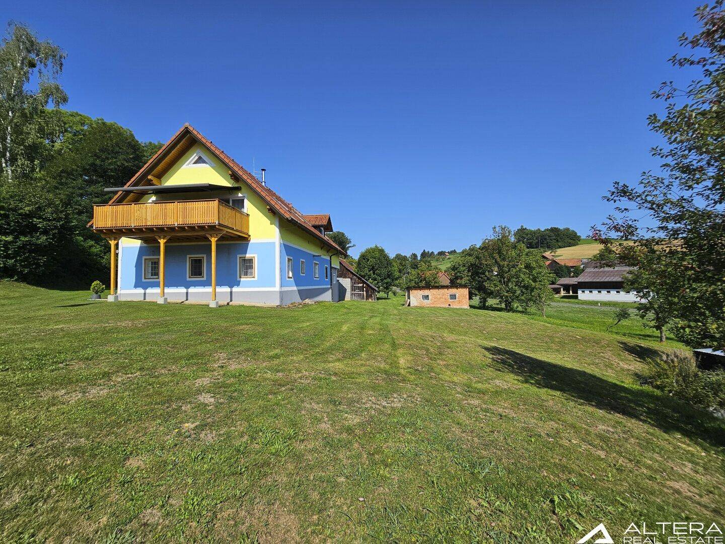 Farbenfrohes Haus mit blau-gelber Fassade, Holzbalkon und weitläufiger grüner Umgebung.