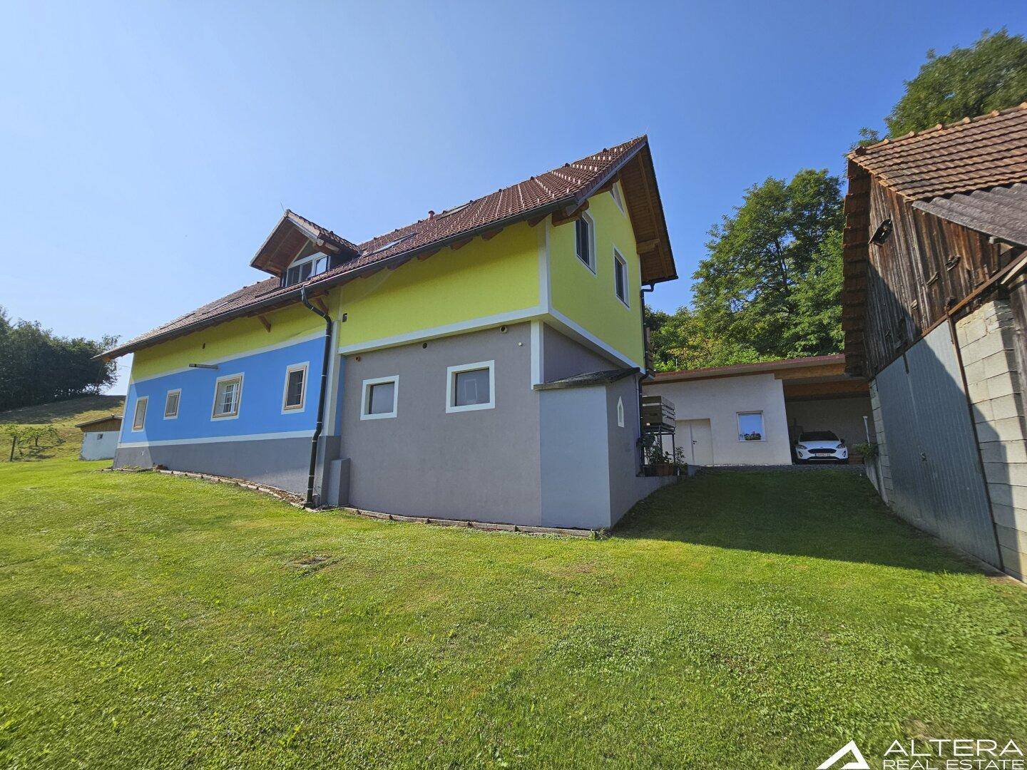 Rückansicht des Hauses mit gelb-blauer Fassade, Carport und angrenzendem Nebengebäude.