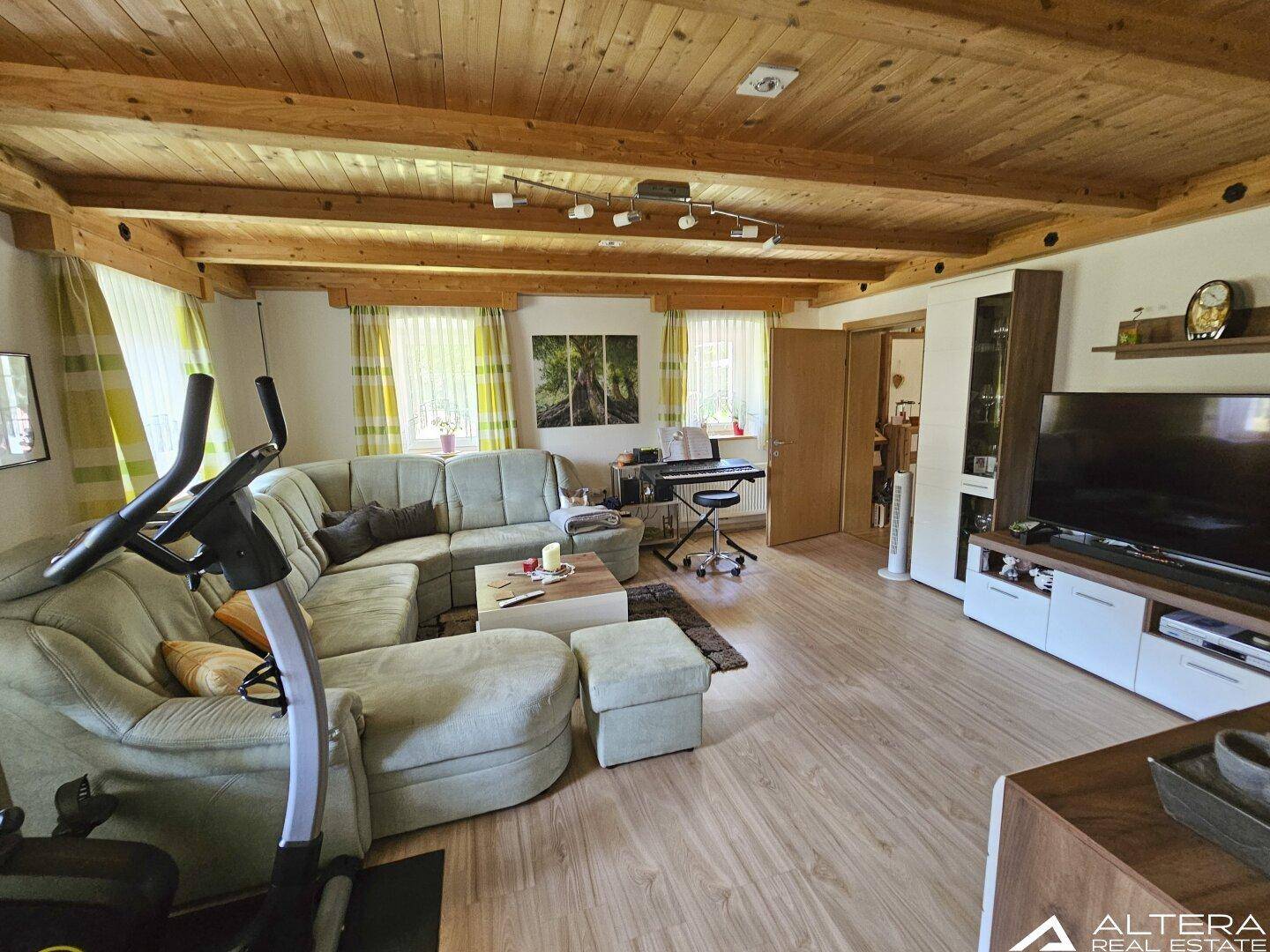 Geräumiges Wohnzimmer mit Fitnessgerät, Ecksofa und Holzbalkendecke, ideal zum Entspannen.