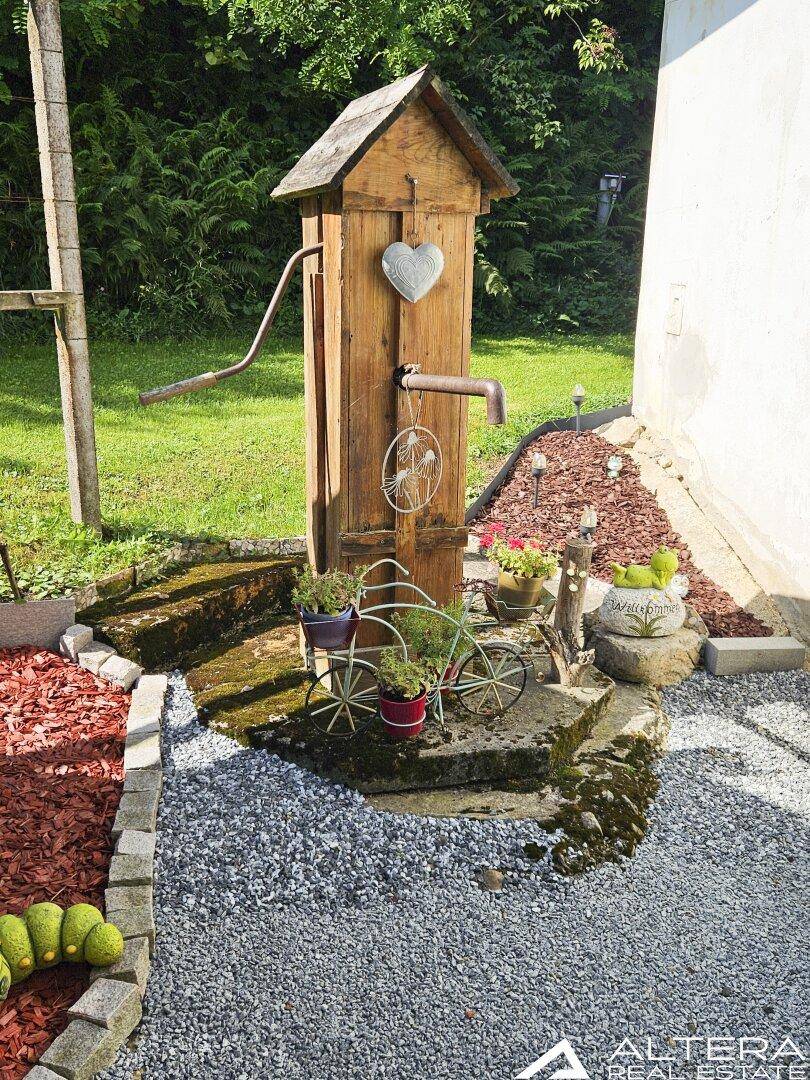 Dekorativer Gartenbereich mit Holzzierbrunnen und Kiesbeet, umgeben von Grünflächen.