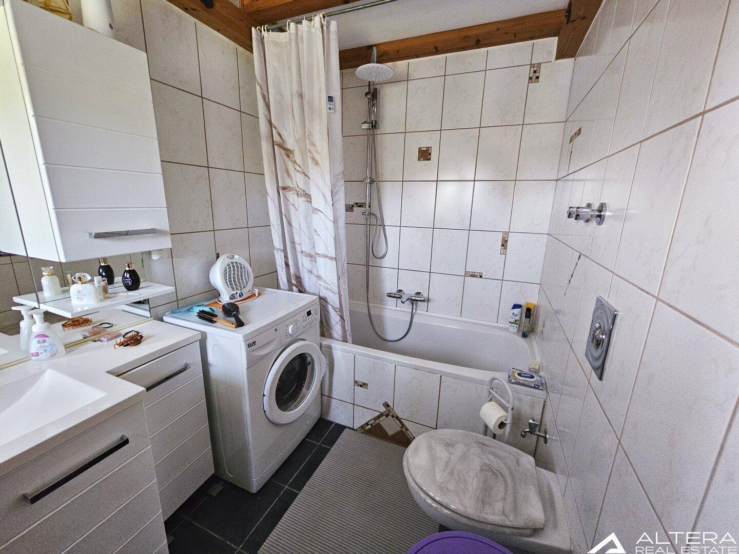 Badezimmer mit Badewanne, Dusche und Waschmaschine, hell gefliest und funktional eingerichtet.